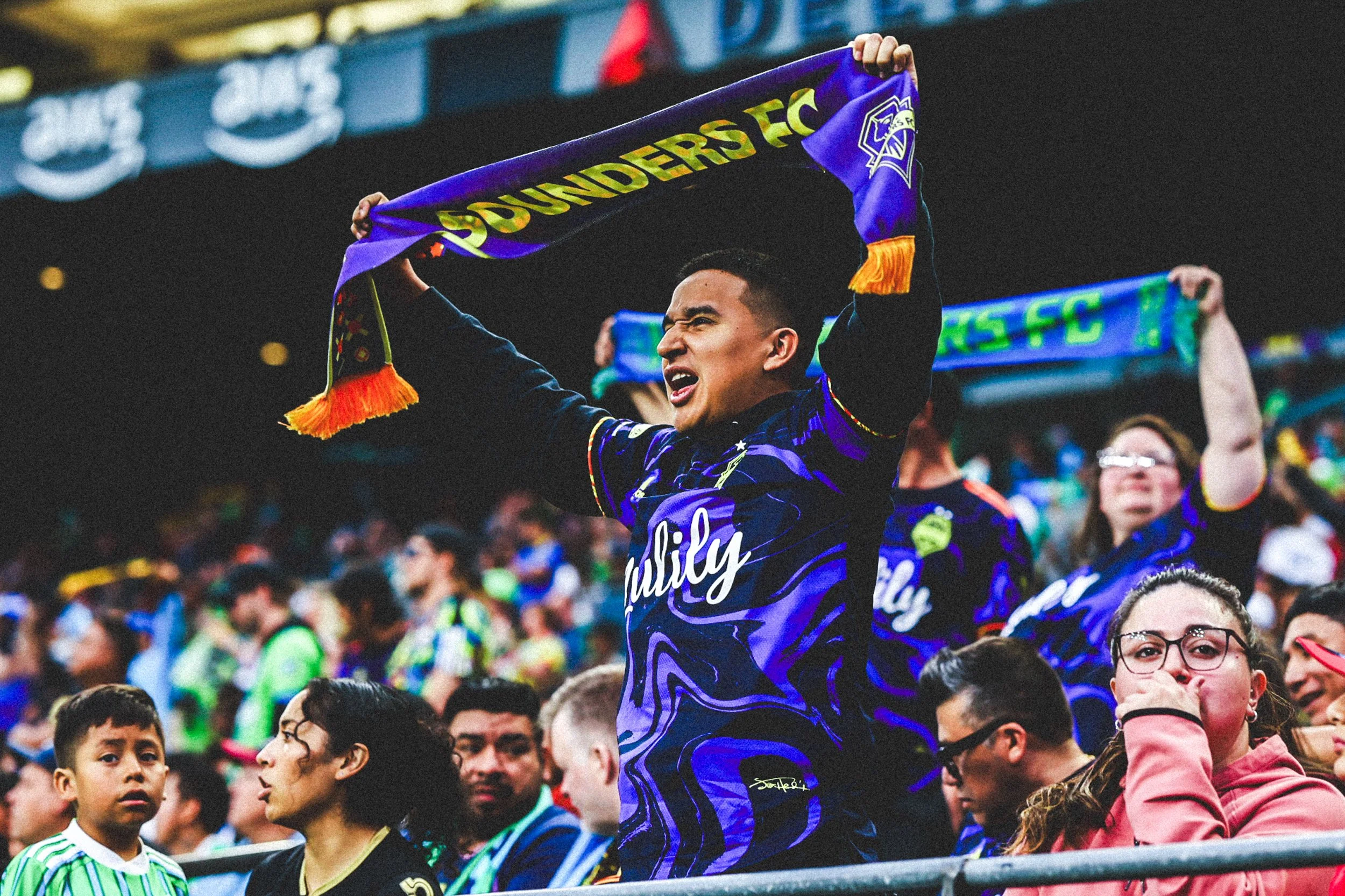 A group of diverse soccer fans at a stadium, with one man in the center holding a purple and yellow scarf above his head, cheering. The spectators are engaged and look excited during the game.