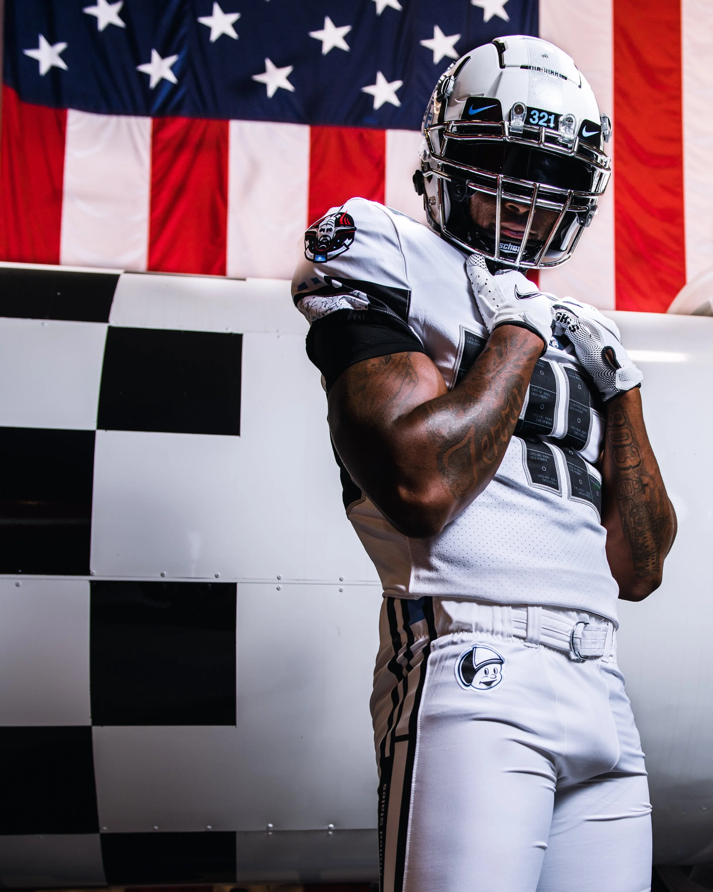 An American football player in uniform stands in front of an aircraft with a checkered pattern, with an American flag in the background.