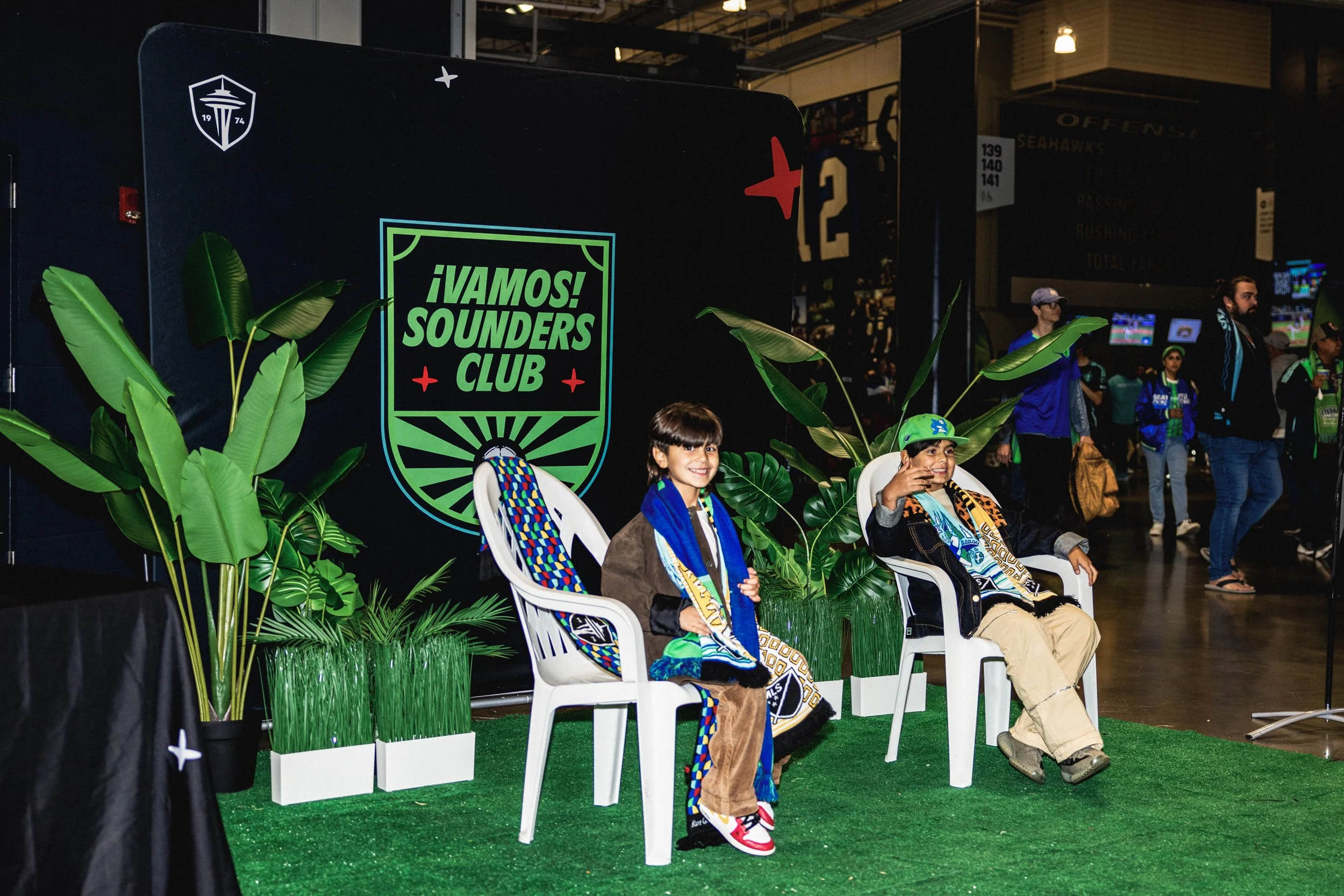 Two children sitting on white chairs in front of a backdrop with the Seahawks logo and green plants, during an event at a Seahawks game.
