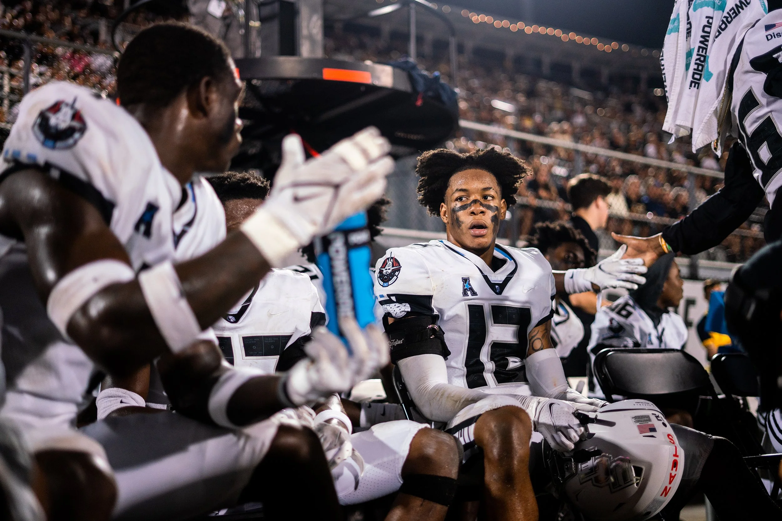 American football players on the sidelines during a game, with one player sitting and looking towards the field, wearing a white jersey with the number 15, surrounded by teammates and a coach in a stadium filled with spectators.