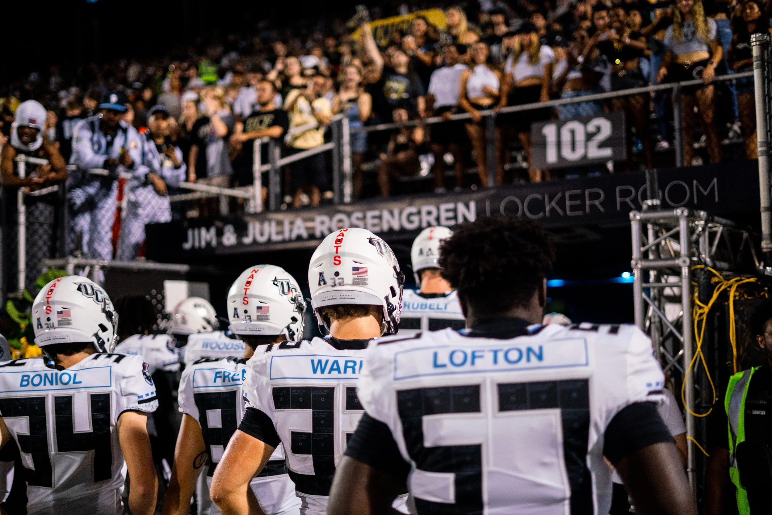 American football team standing on the field with their backs to the camera while fans watch from the stands at night.