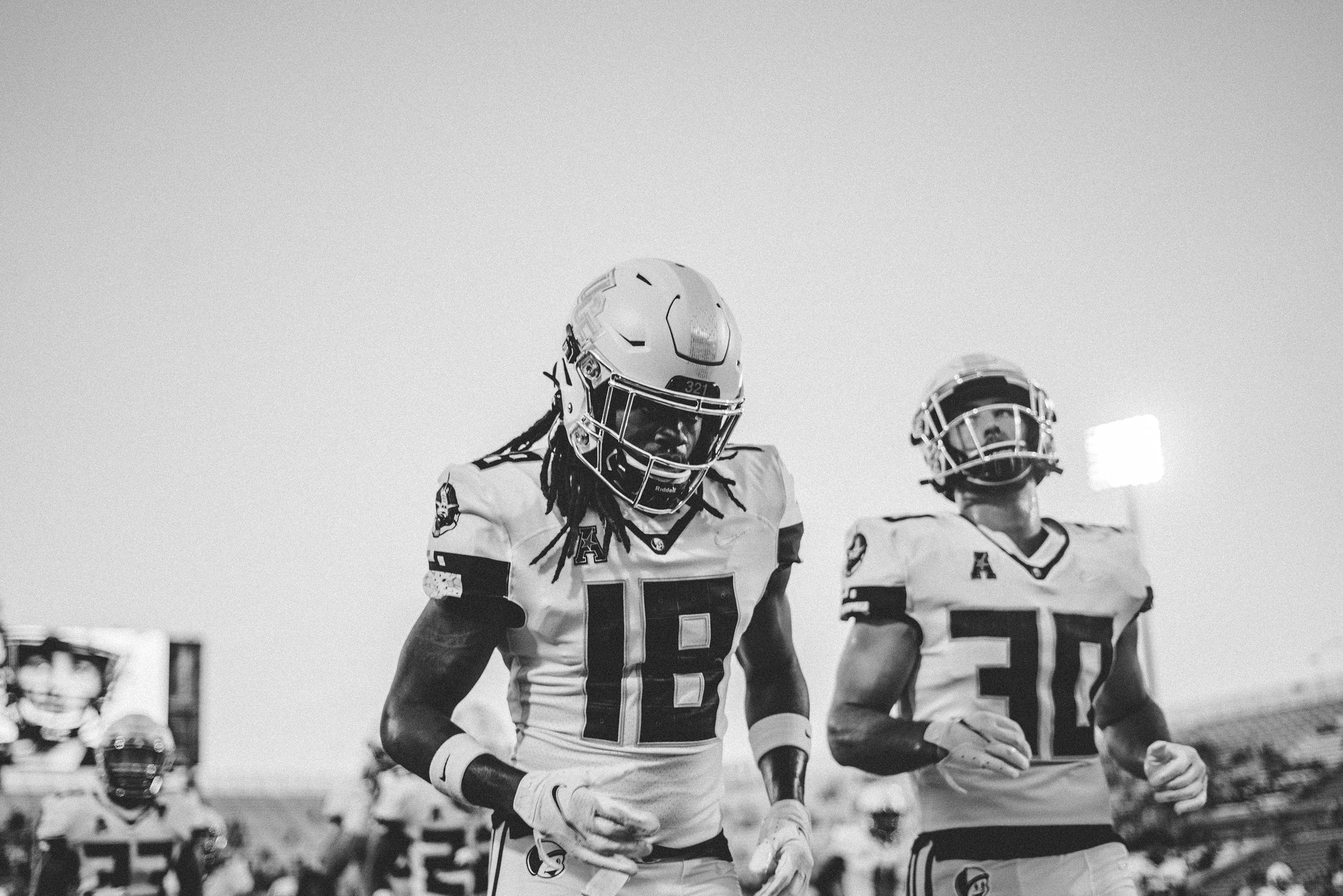 Two football players in uniform, wearing helmets, walking on the field during a game.