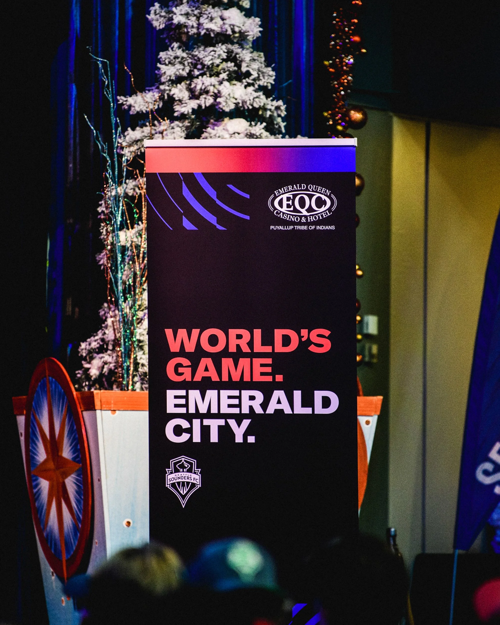 A promotional sign for a sporting event or celebration in Emerald City, featuring the Seattle Sounders FC logo, with a Christmas tree decorated with ornaments and a toy cannon nearby.