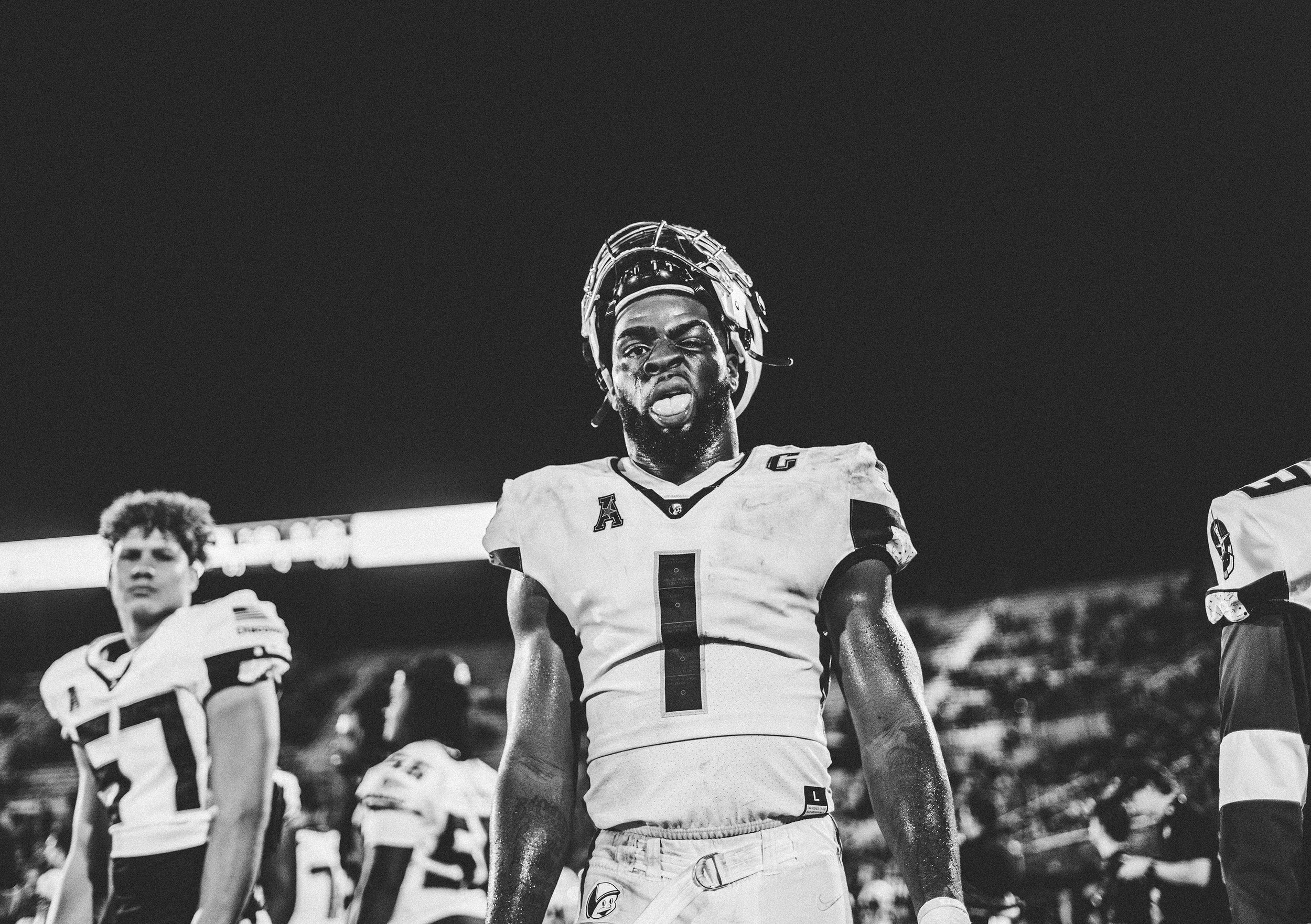 Black and white photograph of American football players on the field during a game, with one player in the center making an intense facial expression, sticking his tongue out and wearing a helmet. Other players are visible in the background.