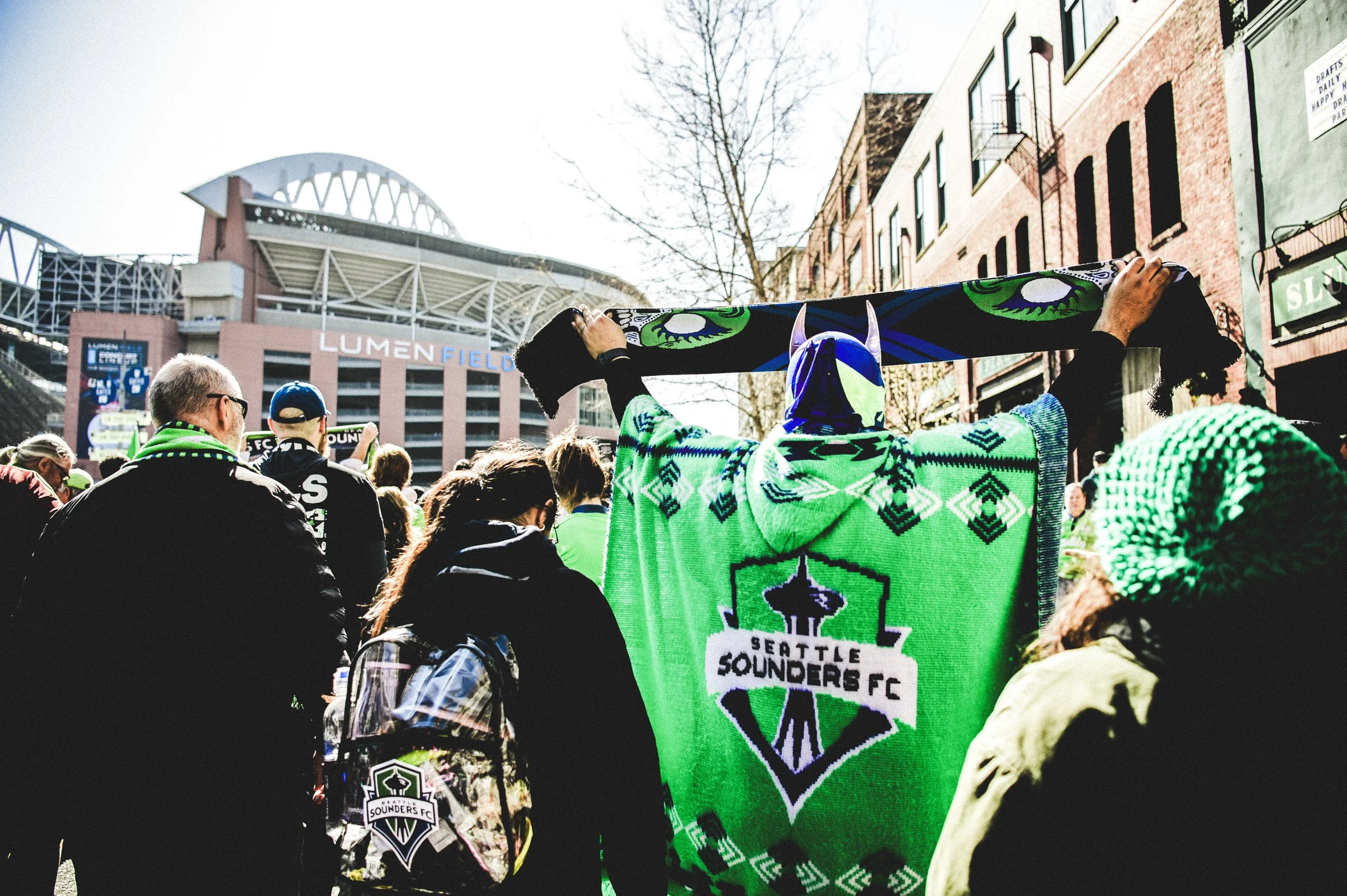 Crowd of Seattle Sounders FC fans gathered outdoors near Lumen Field stadium, some holding a large green Seahawks-themed scarf and wearing team merchandise, during daytime.