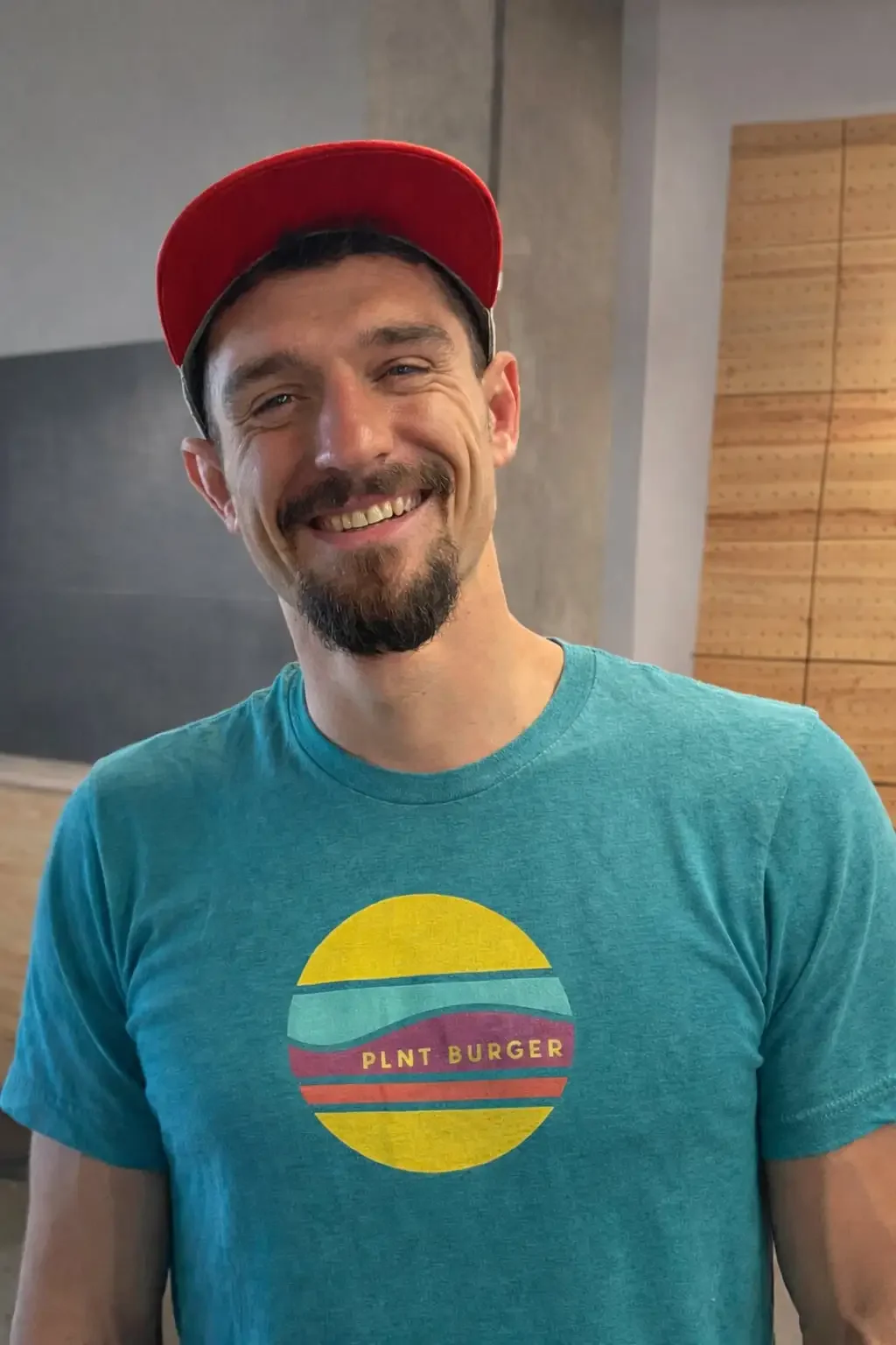 A man with a dark beard and mustache, wearing a red cap and a turquoise PLNT Burger t-shirt, smiles at the camera in an indoor setting with neutral-colored walls.