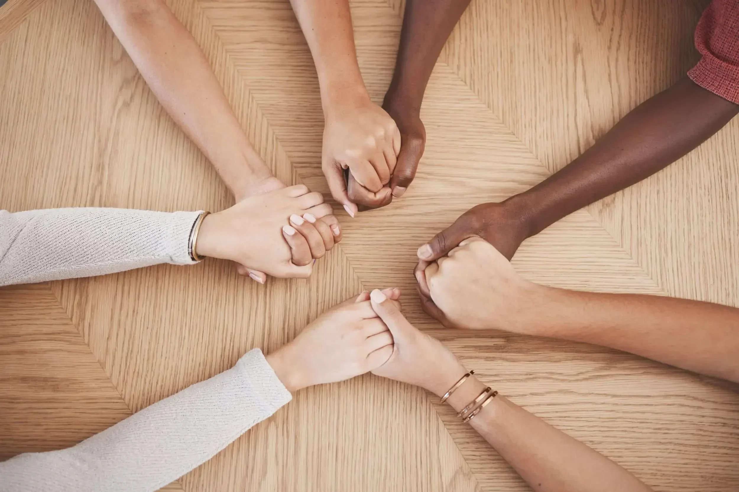 Six diverse hands holding each other in a supportive circle on a wooden table, symbolizing unity and connection.