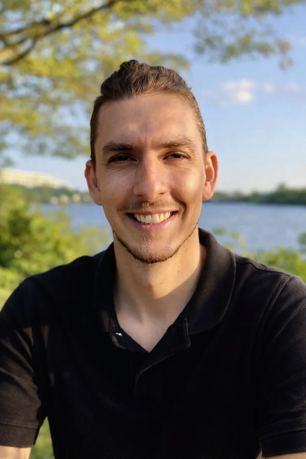 A man with brown hair tied back, wearing a black polo shirt, smiles while sitting outdoors near a river with green trees and blue sky in the background.