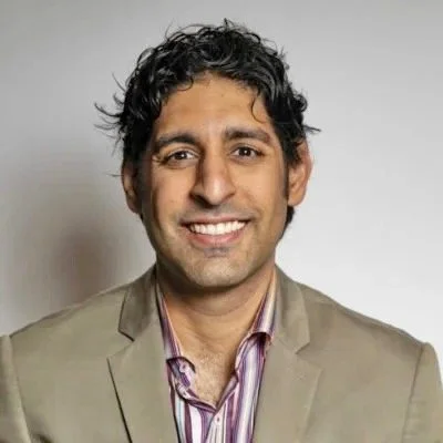 A man with dark, curly hair and medium skin tone smiles at the camera. He is wearing a beige blazer over a striped shirt, standing in front of a plain light background.