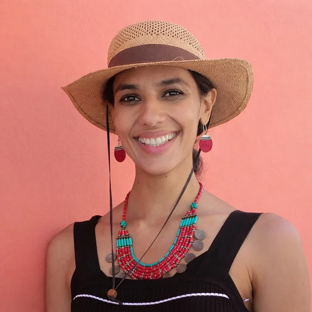 A woman wearing a straw hat, red earrings, and a colorful beaded necklace smiles in front of a pinkish-orange wall. She is dressed in a sleeveless black top with thin straps.