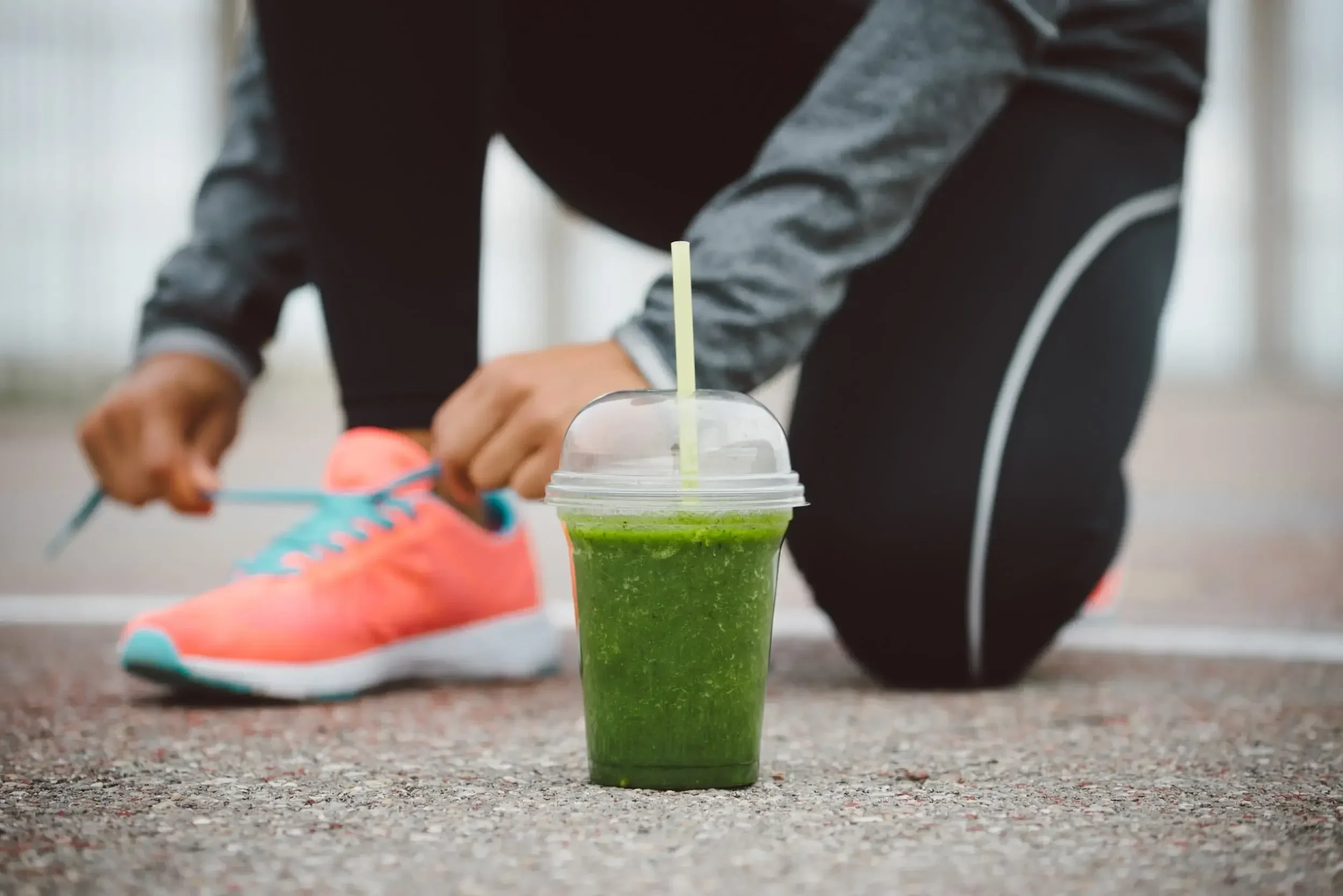 A person in workout gear ties shoelaces near a green smoothie in a plastic cup with a straw on the ground.
