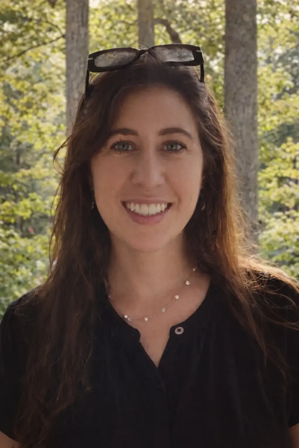 A woman with long brown hair, wearing a black top, a beaded necklace, and glasses resting on her head, smiles while standing outdoors with green trees in the background.