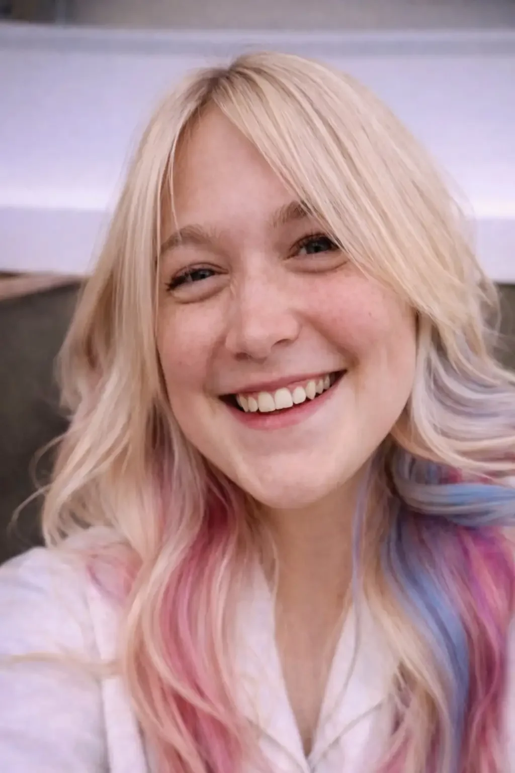 A young person with long, wavy blonde hair featuring pastel pink and blue streaks smiles at the camera. They are wearing a light-colored top and the background is blurred.