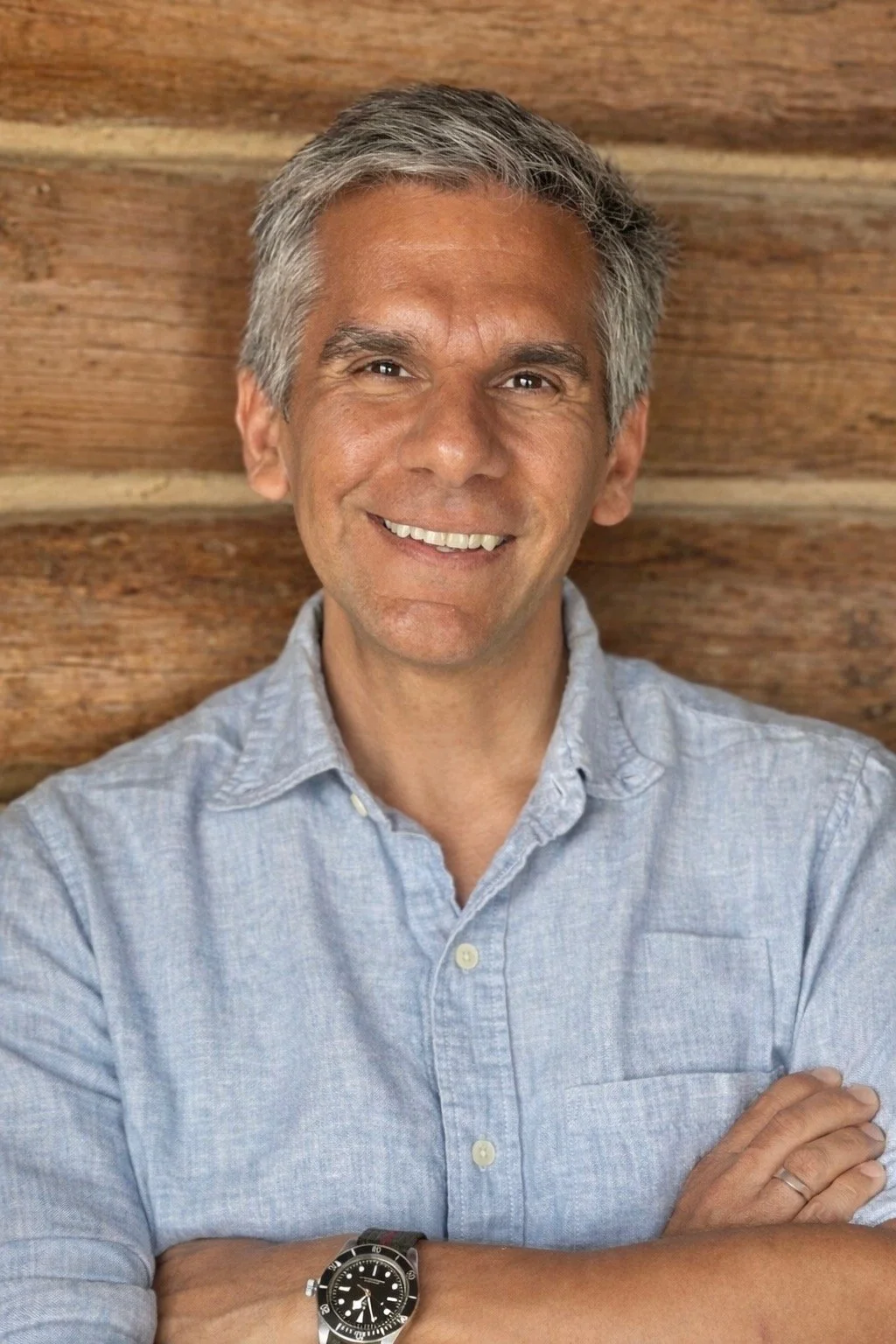 A middle-aged man with gray hair smiles, arms crossed, wearing a light blue button-up shirt and a wristwatch, standing in front of a wooden wall.
