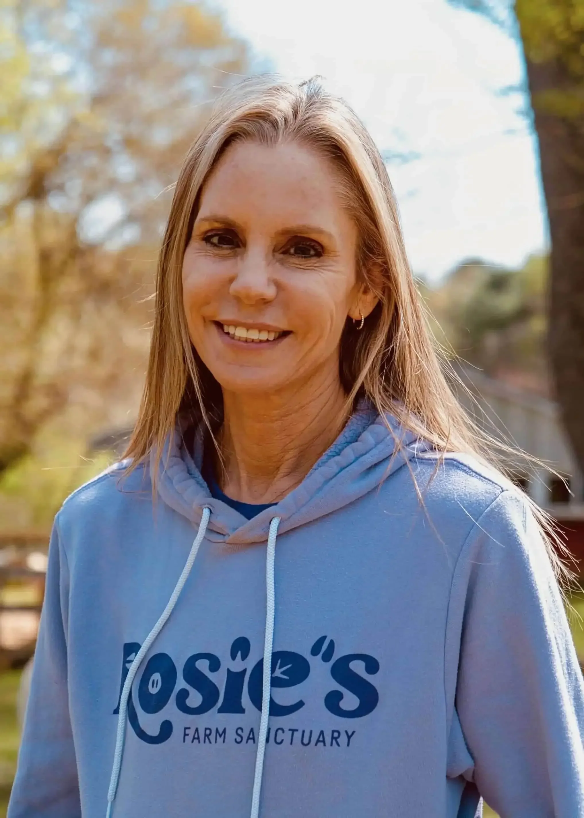 A woman with long blonde hair, wearing a light blue hoodie that says "Rosie's Farm Sanctuary," stands outdoors smiling, with blurred trees and sunlight in the background.