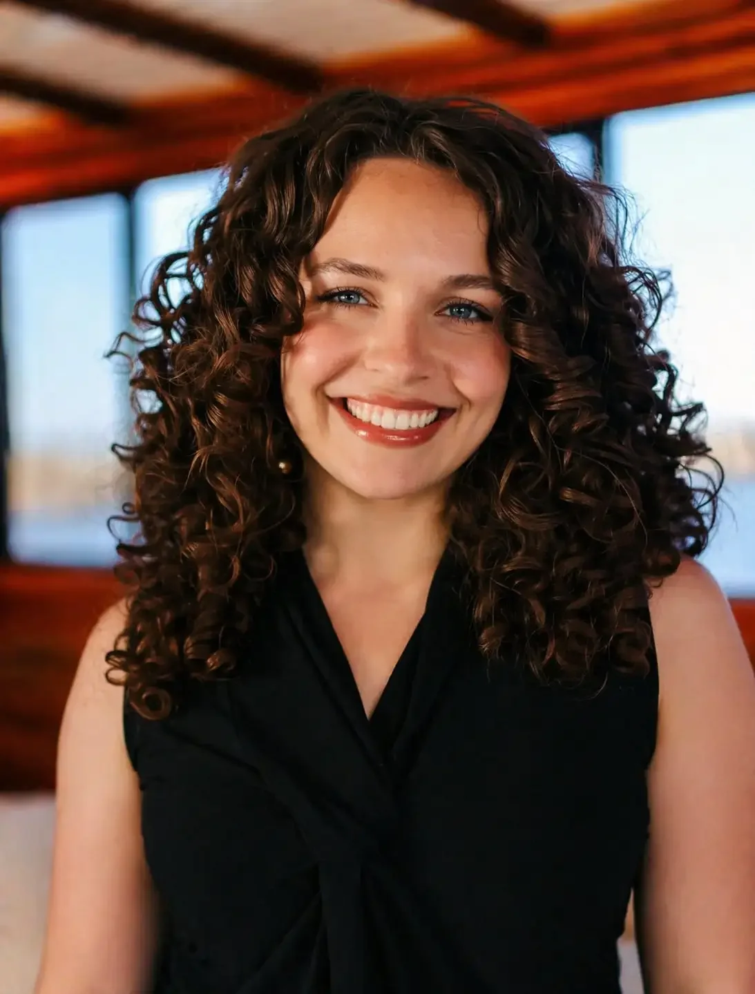 A woman with curly brown hair and blue eyes smiles warmly at the camera. She is wearing a sleeveless black top and stands indoors, with large windows and natural light in the background.