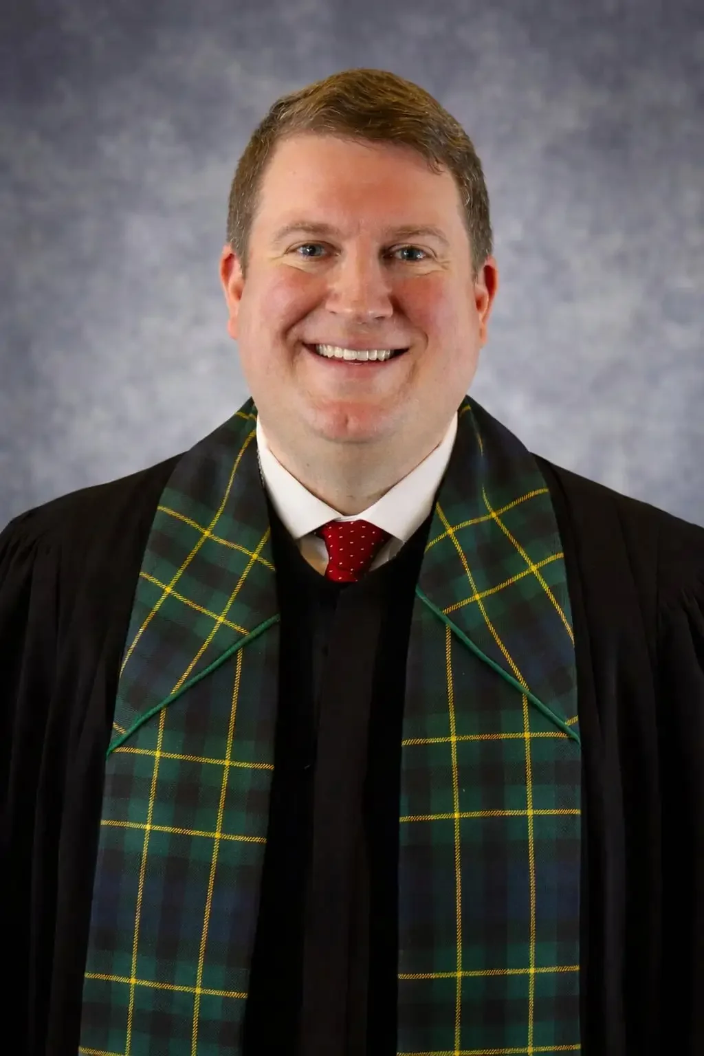 A smiling man in a black robe with a green and yellow plaid stole and a red polka dot tie stands in front of a mottled gray backdrop.