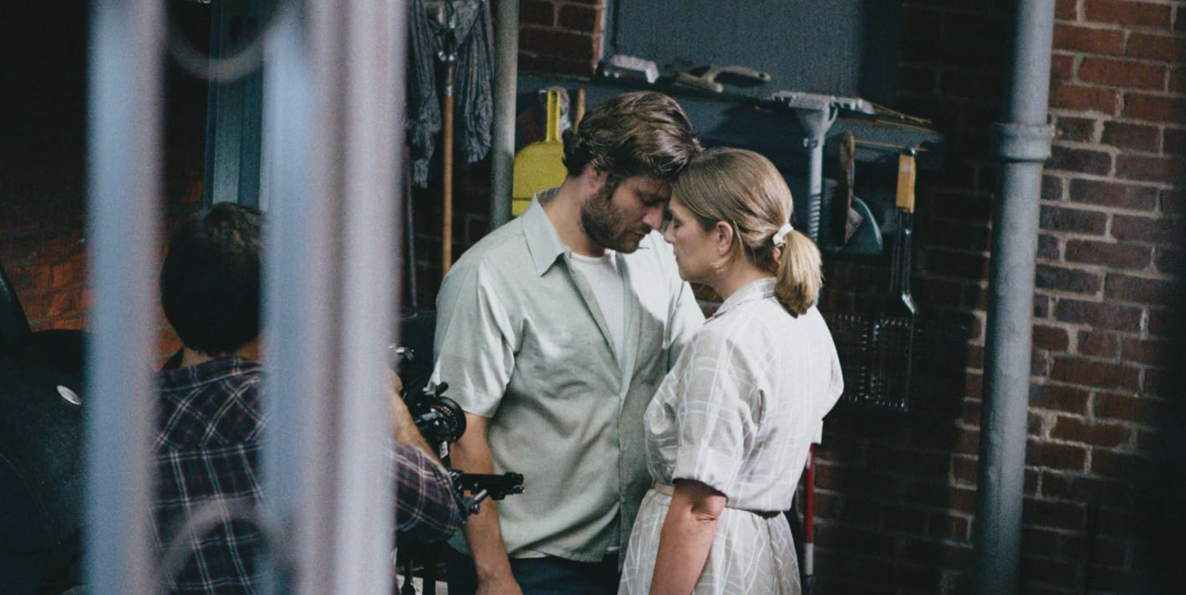 A scene from the movie 'The Fault in Our Stars' showing a young man and woman with foreheads touching, looking into each other's eyes in a garage or basement setting surrounded by brick walls and various tools.