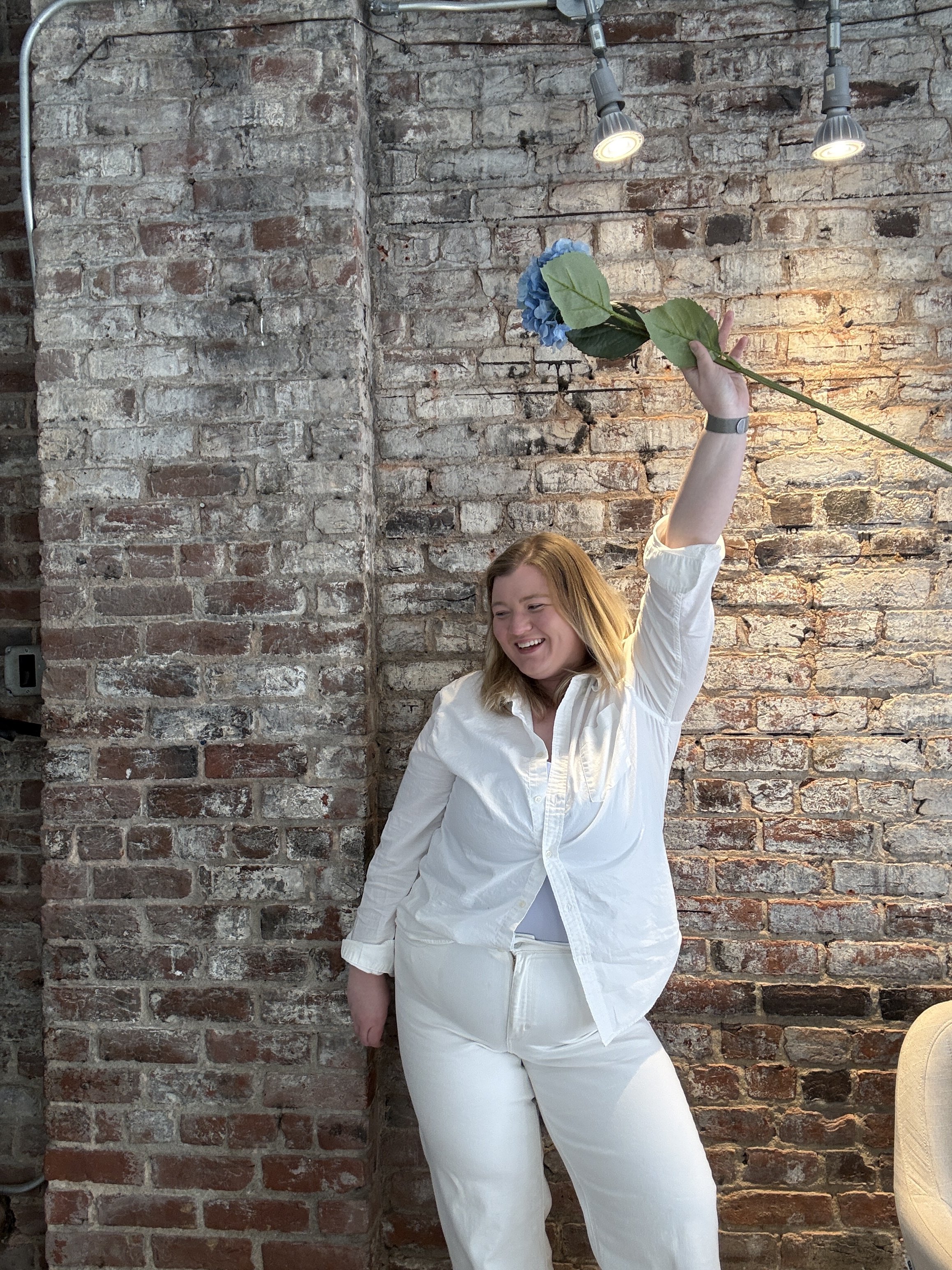 A woman in an all-white outfit smiling and raising her left arm holding a blue flower with large green leaves in front of a brick wall.