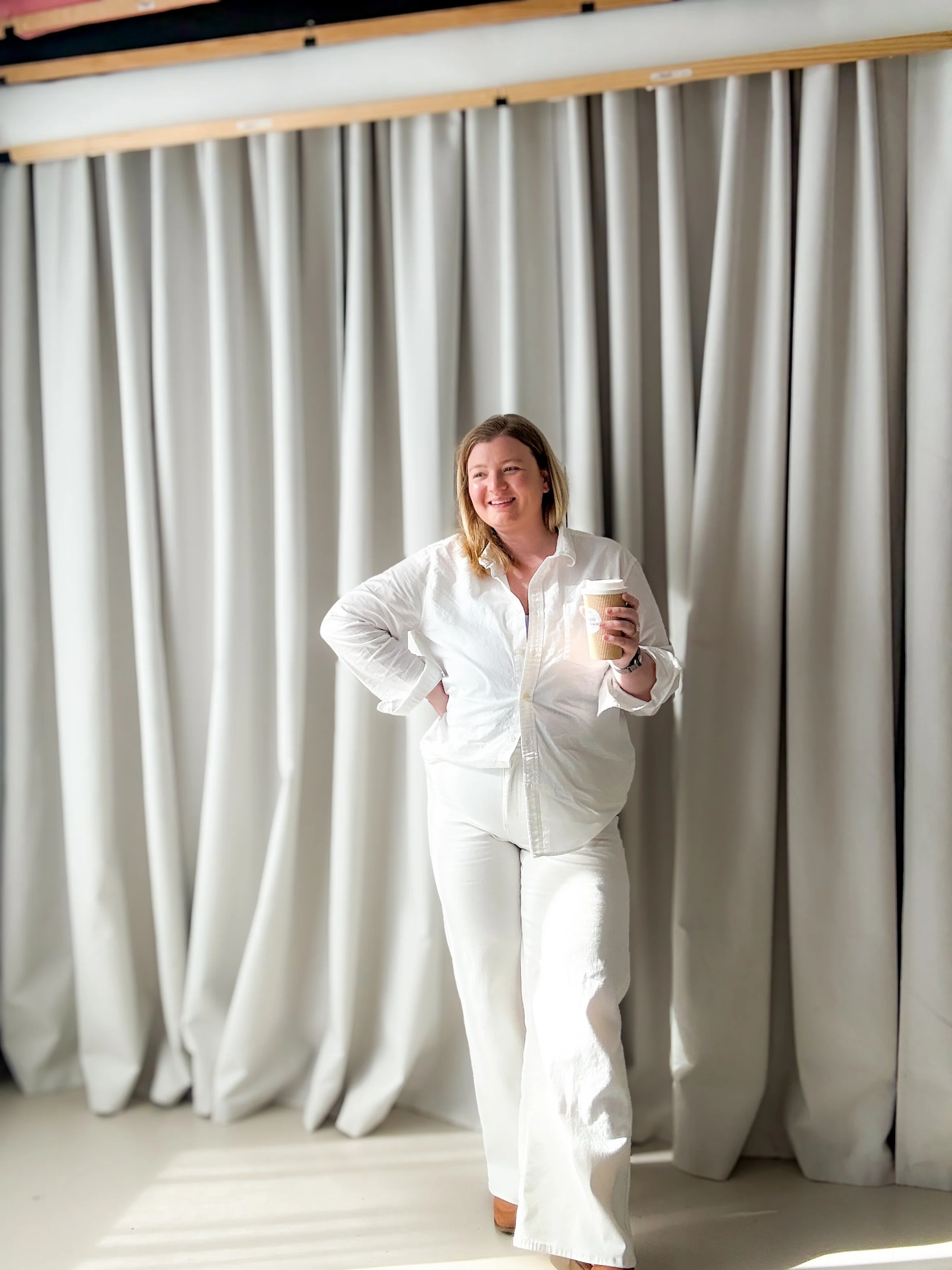 A woman in white clothing holding a coffee cup standing in front of cream-colored curtains.