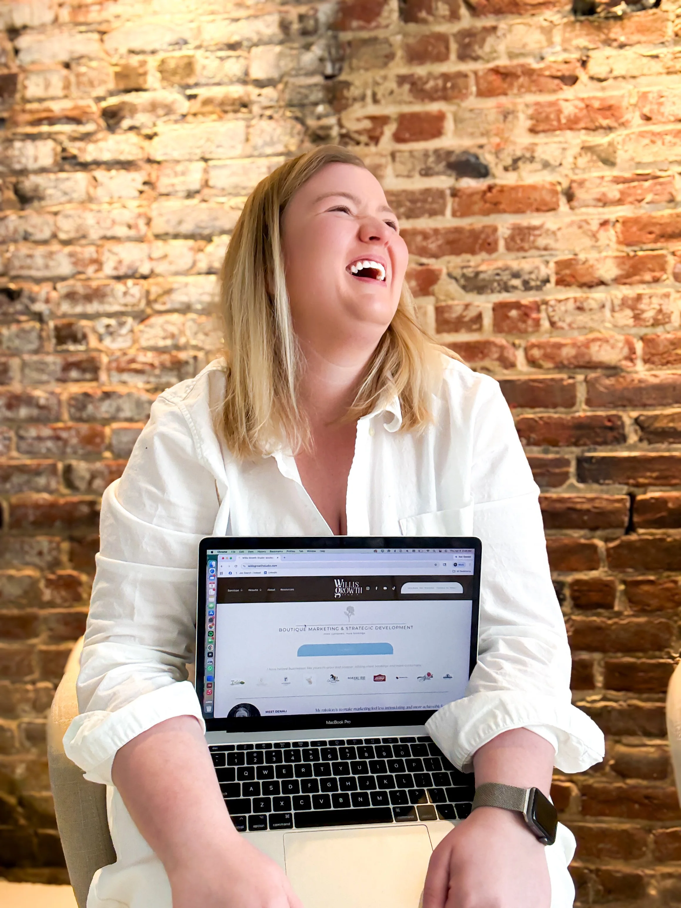 A woman with blonde hair wearing a white shirt is sitting in front of a brick wall, laughing and holding a laptop on her lap. The laptop screen displays a website. She is wearing a black smartwatch on her left wrist.