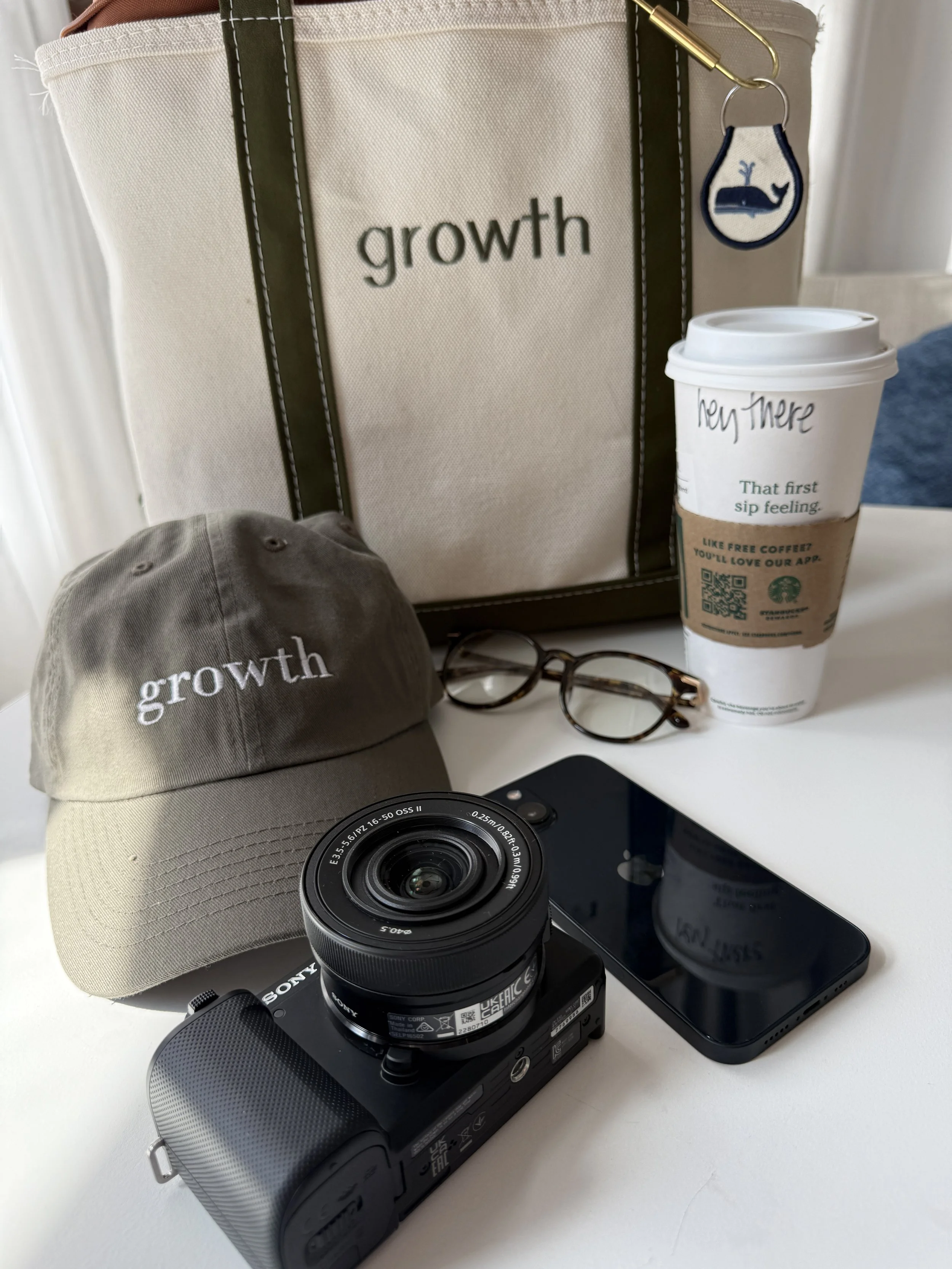 A collection of personal items on a white desk, including a beige cap with the word 'growth' embroidered on it, a black camera, a smartphone, a pair of eyeglasses, a reusable coffee cup with a sleeve, a tote bag with the word 'growth' and a whale keychain attached, and a framed sign with the word 'growth'.