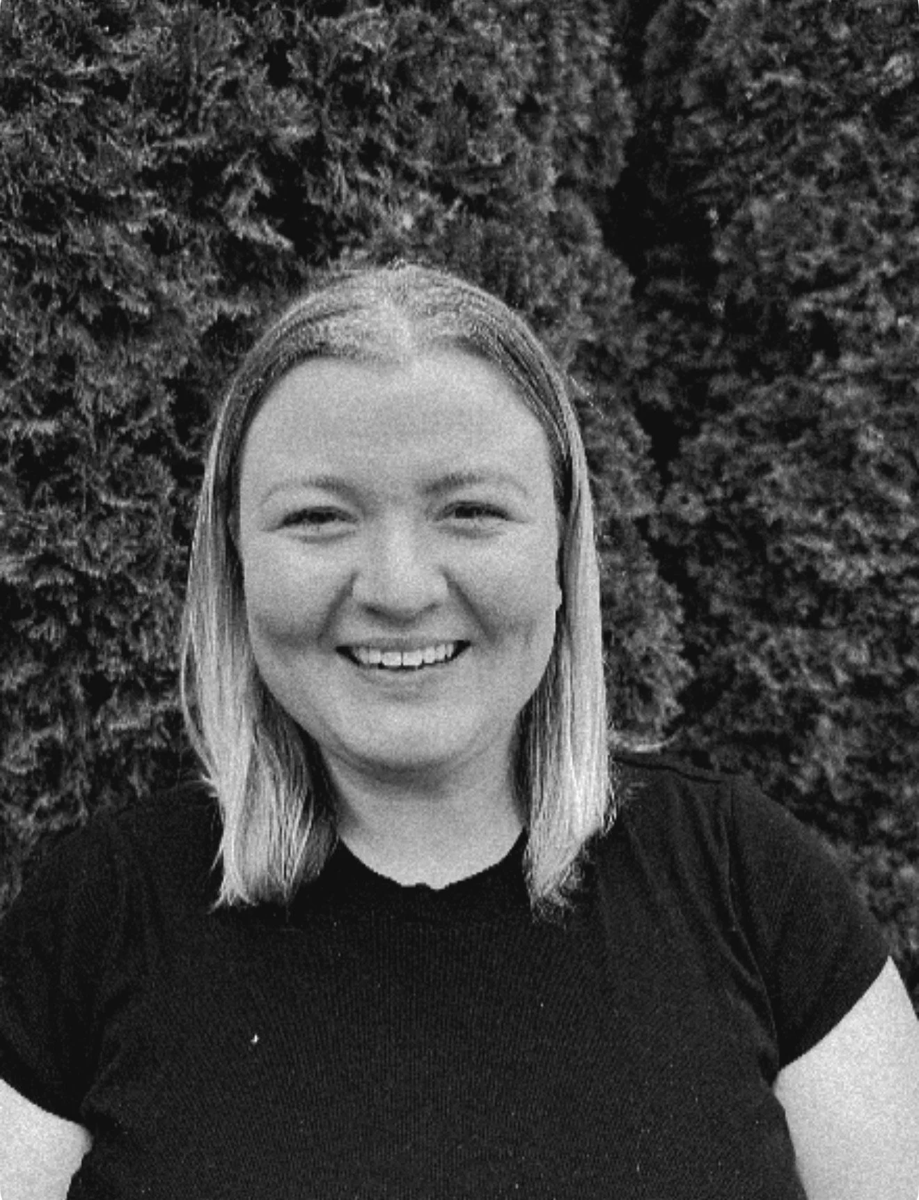 A woman with shoulder-length hair smiling outdoors in front of a dense hedge, wearing a black top.