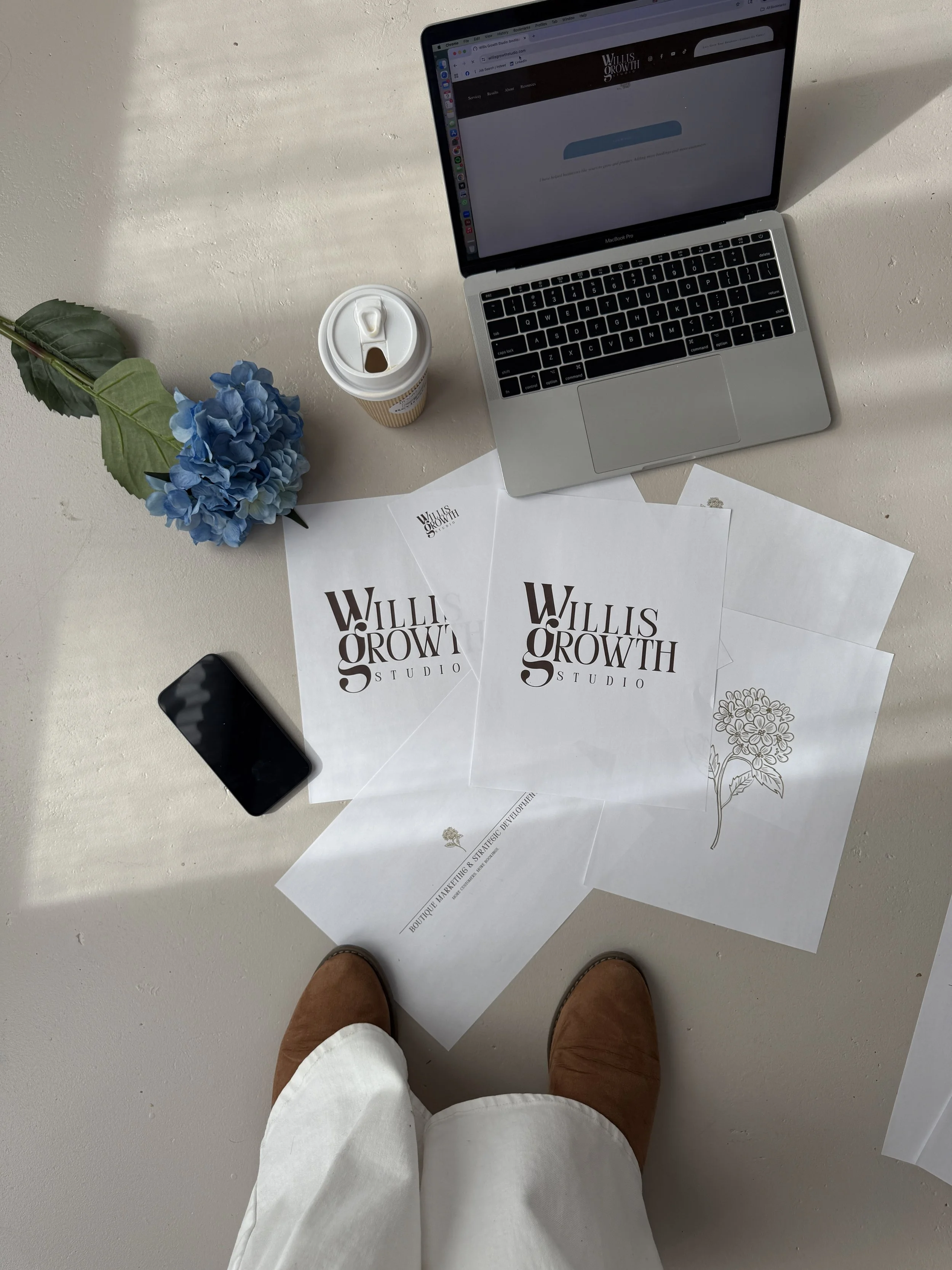 Overhead view of a workspace with a person standing at it, wearing white pants and brown shoes. The workspace includes a MacBook laptop, a black smartphone, a coffee cup, a blue hydrangea flower, and several sheets of paper with 'Willis Growth Studio' printed on them, along with a hand-drawn flower illustration.