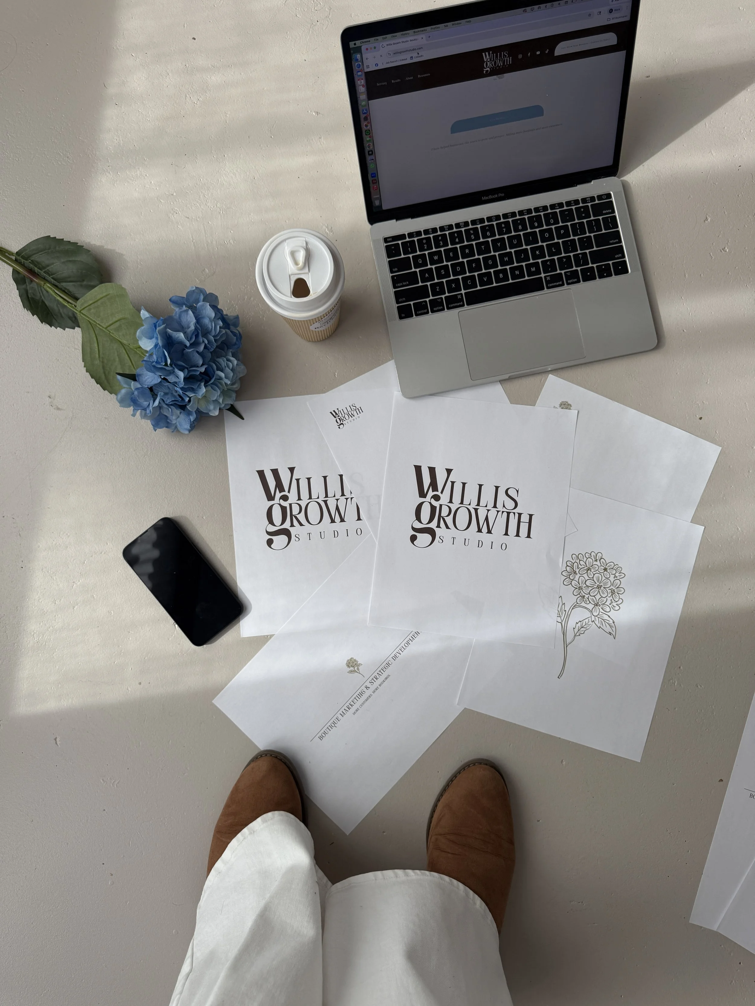 Desk with a MacBook, a coffee cup, a smartphone, a blue flower, and printed papers with 'WILLIS GROWTH STUDIO' logos.