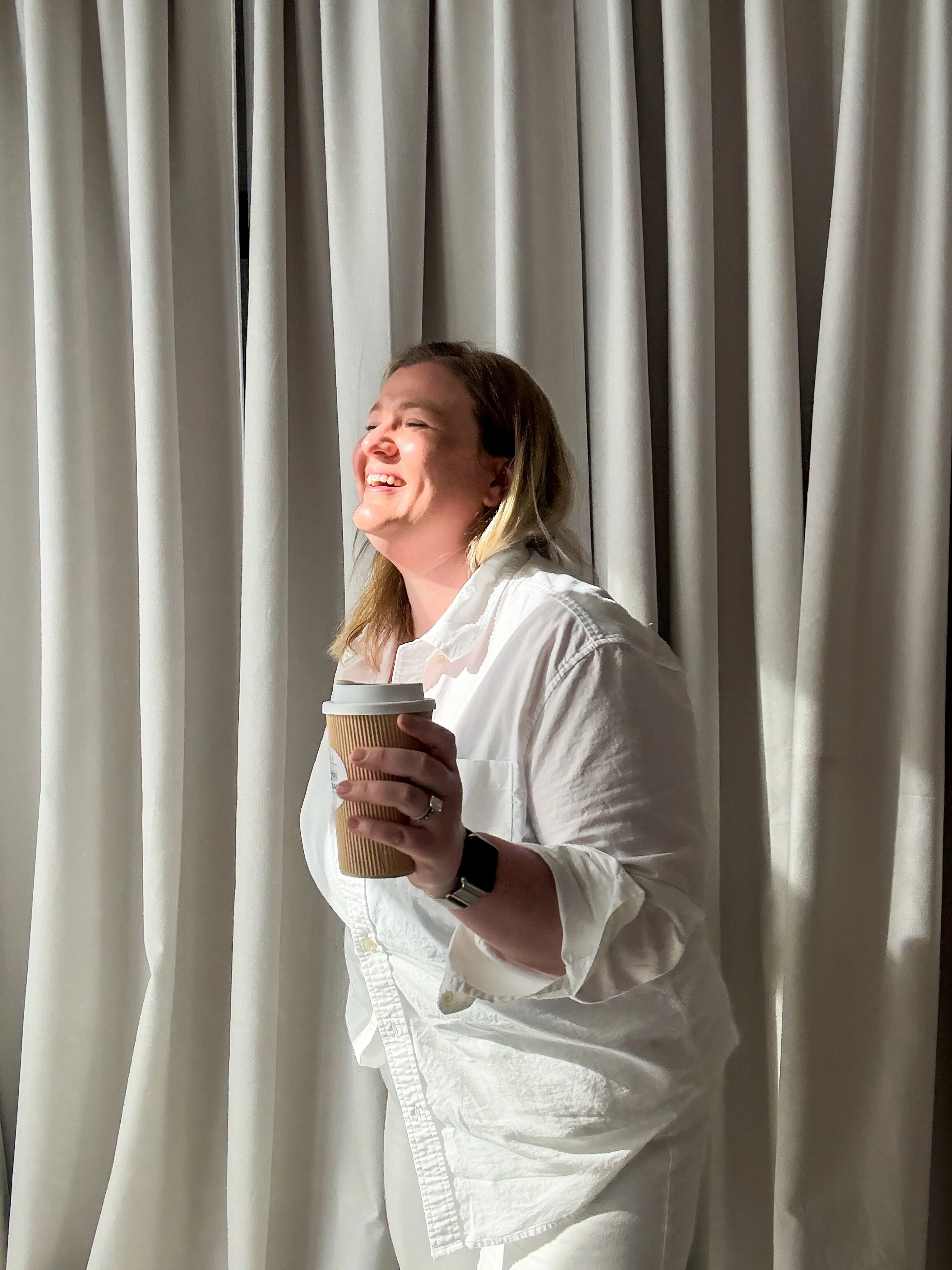 Woman with shoulder-length blonde hair, smiling with eyes closed, holding a to-go coffee cup in front of beige curtains.