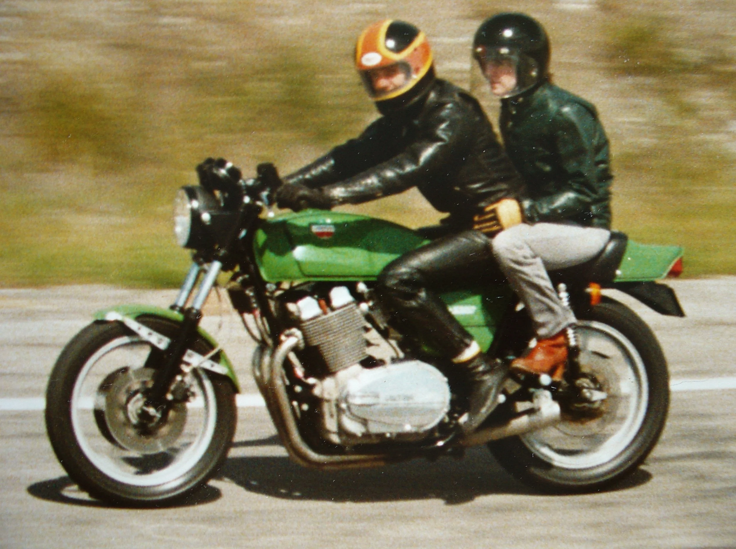 Two people riding a green motorcycle on the road, wearing black leather jackets and black helmets, with the passenger holding onto the rider.
