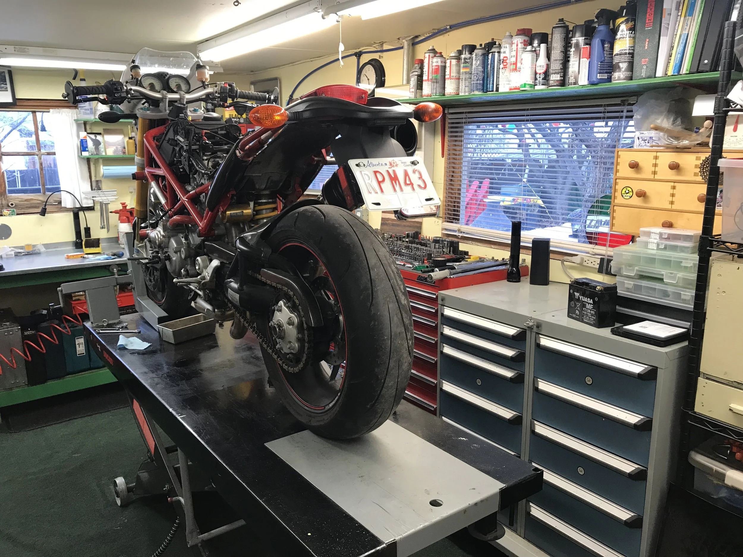 In a garage, a motorcycle is on a workbench with the rear wheel removed. The garage has shelves with spray cans, machines, and tools, with a window showing an outdoor scene.