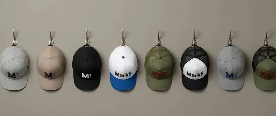 A row of eight baseball caps hanging on hooks against a beige wall, with different colors and logos.