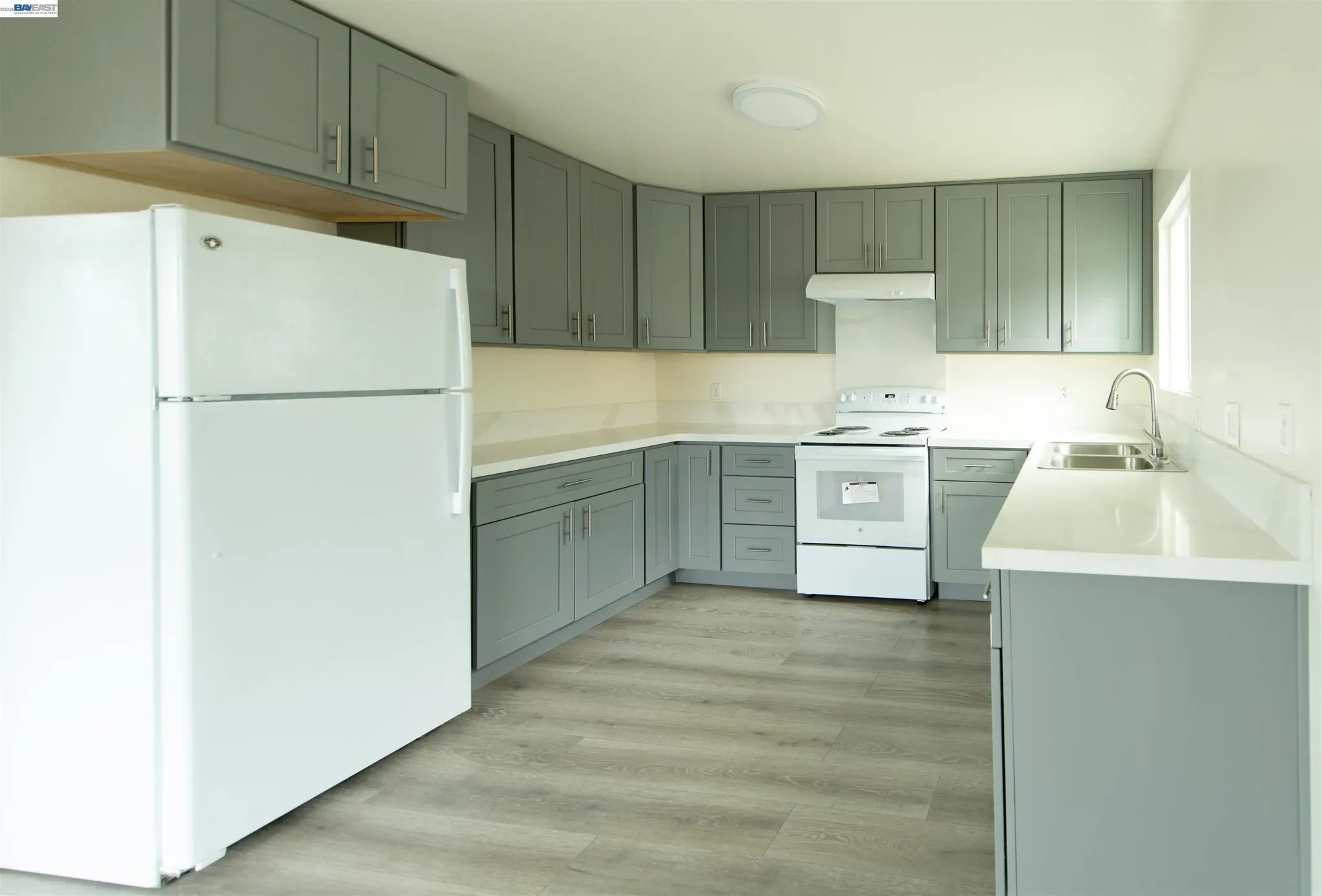 A kitchen with gray cabinets, a white refrigerator, stove, and double sink. The floor is wood and there is a window on the right side.