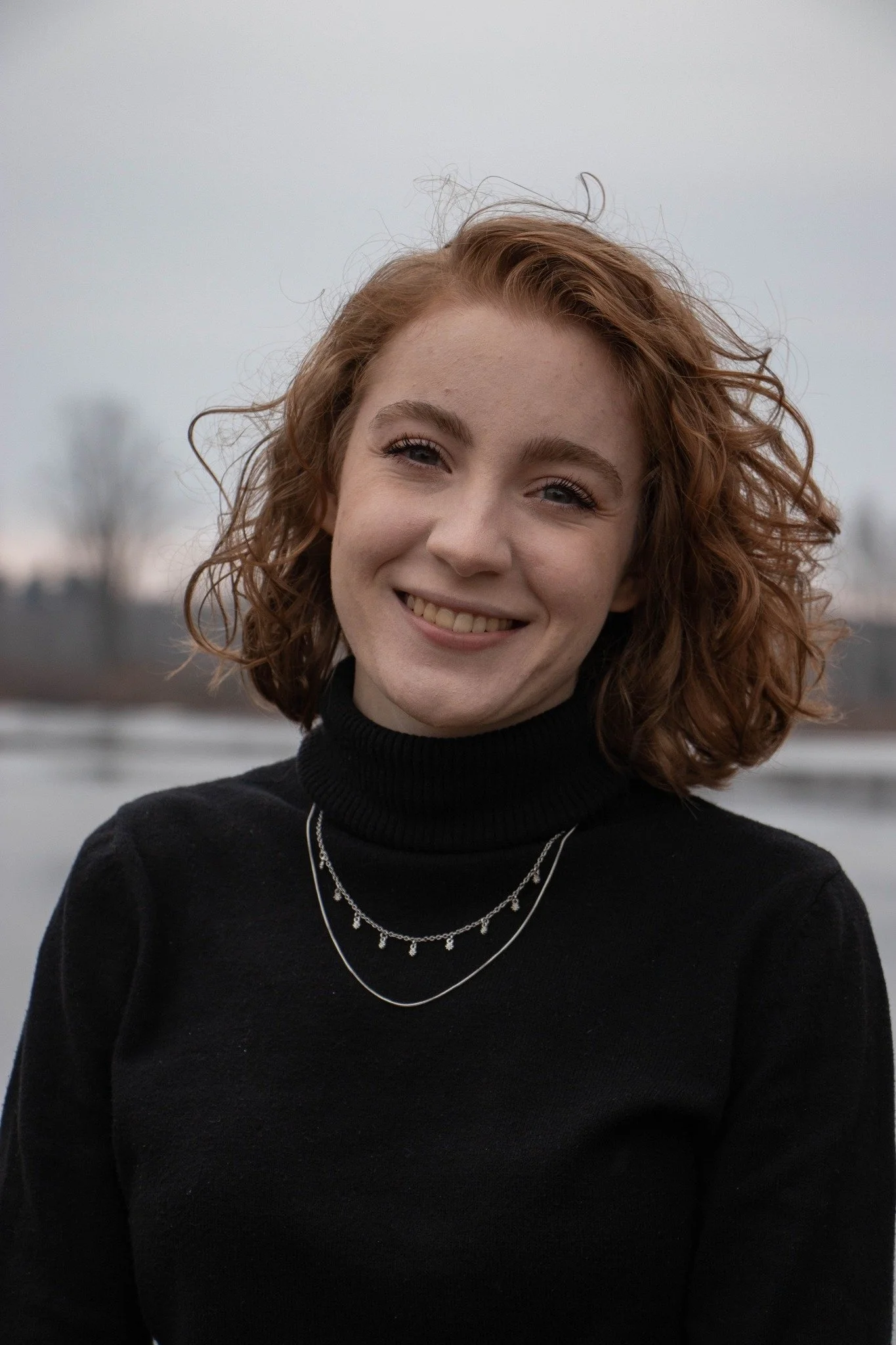 A young woman with curly red hair smiling outdoors by a body of water on a cloudy day, wearing a black turtleneck and layered necklaces.