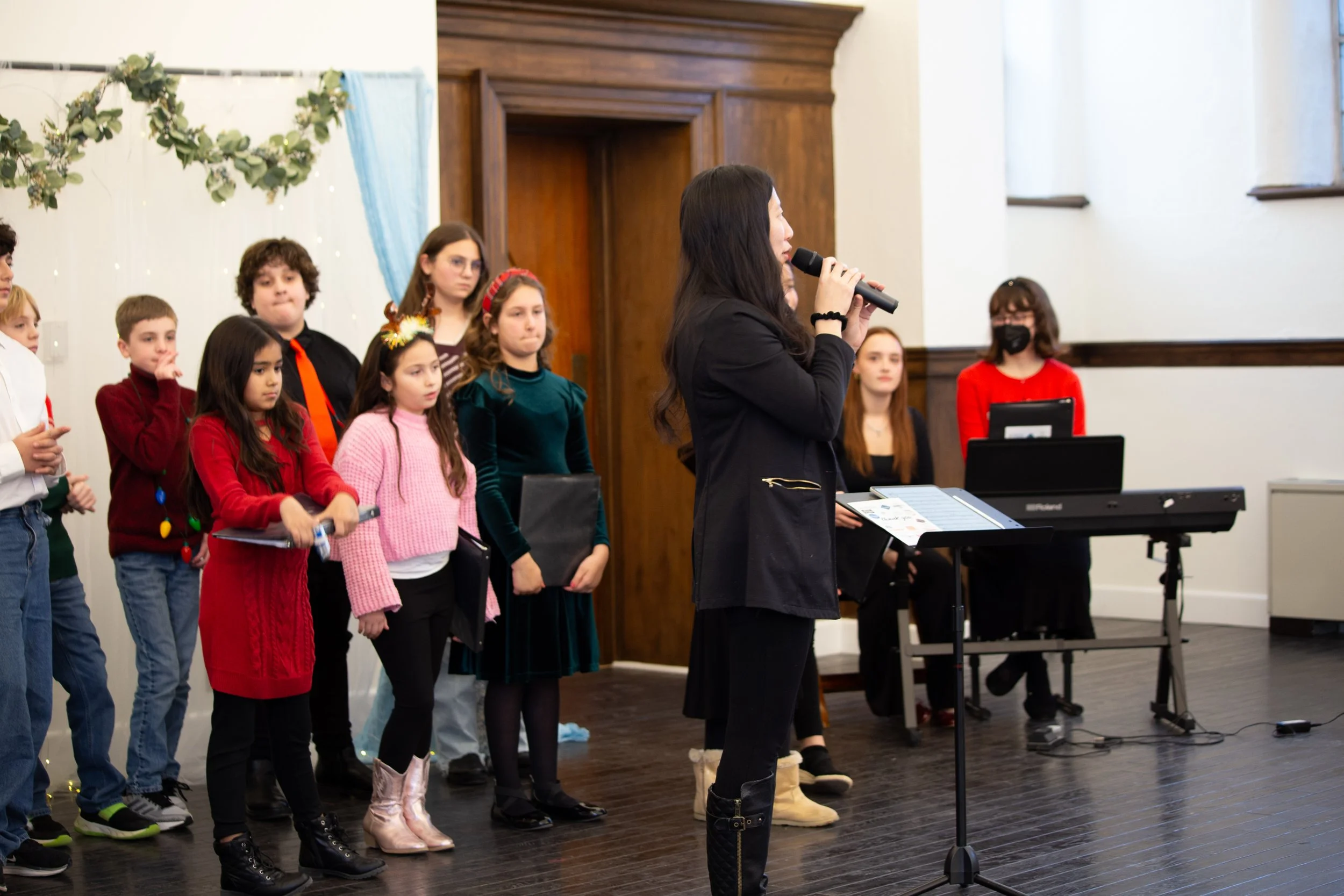 A group of children and a woman with long dark hair, dressed in black, are on stage during a school holiday performance. The children are dressed in festive clothing, with some wearing Christmas sweaters and holding scripts or microphones. A woman is