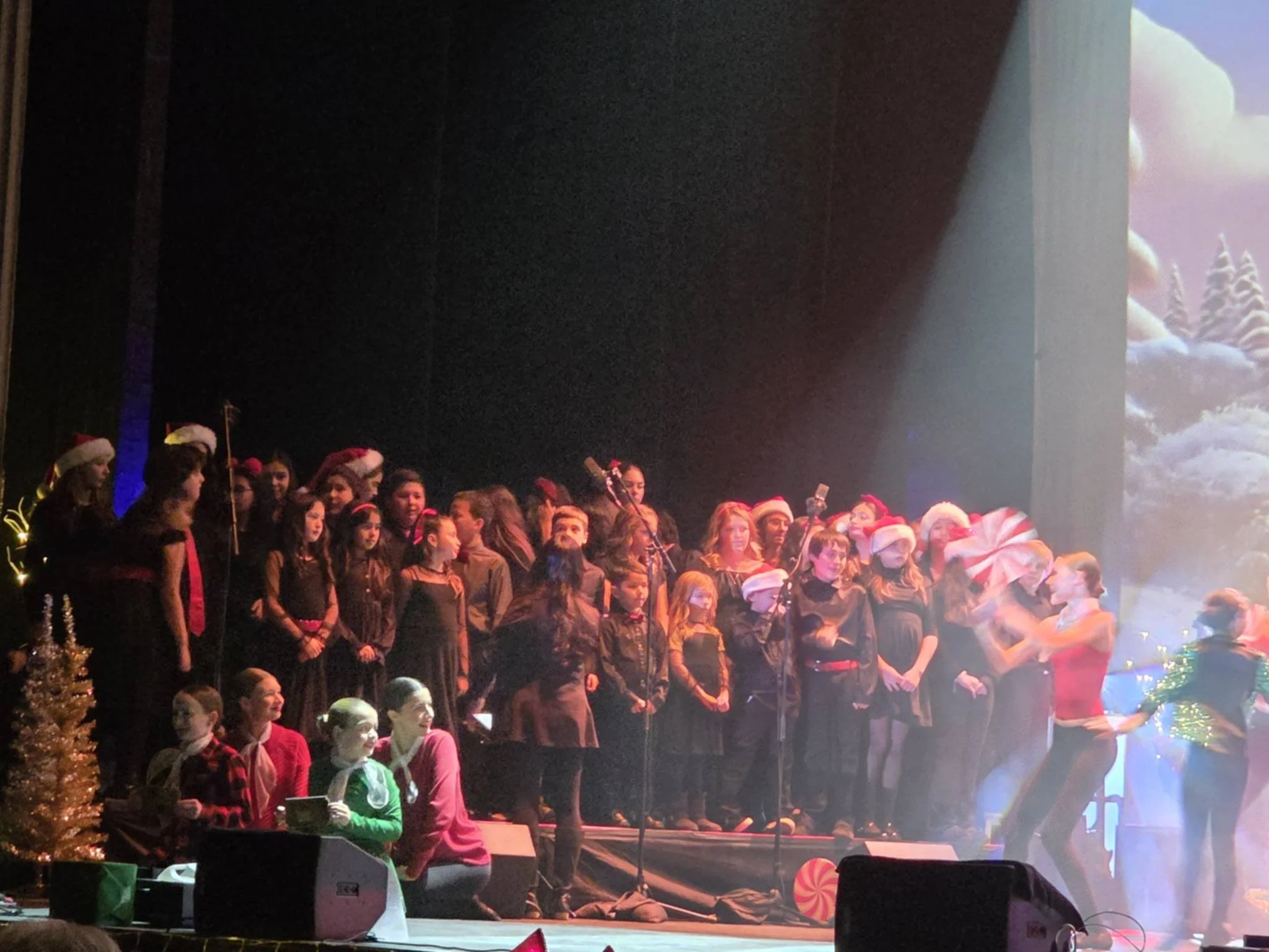 Children and adults on stage performing Christmas carol show, many wearing Santa hats, with festive decorations and a winter scene backdrop.
