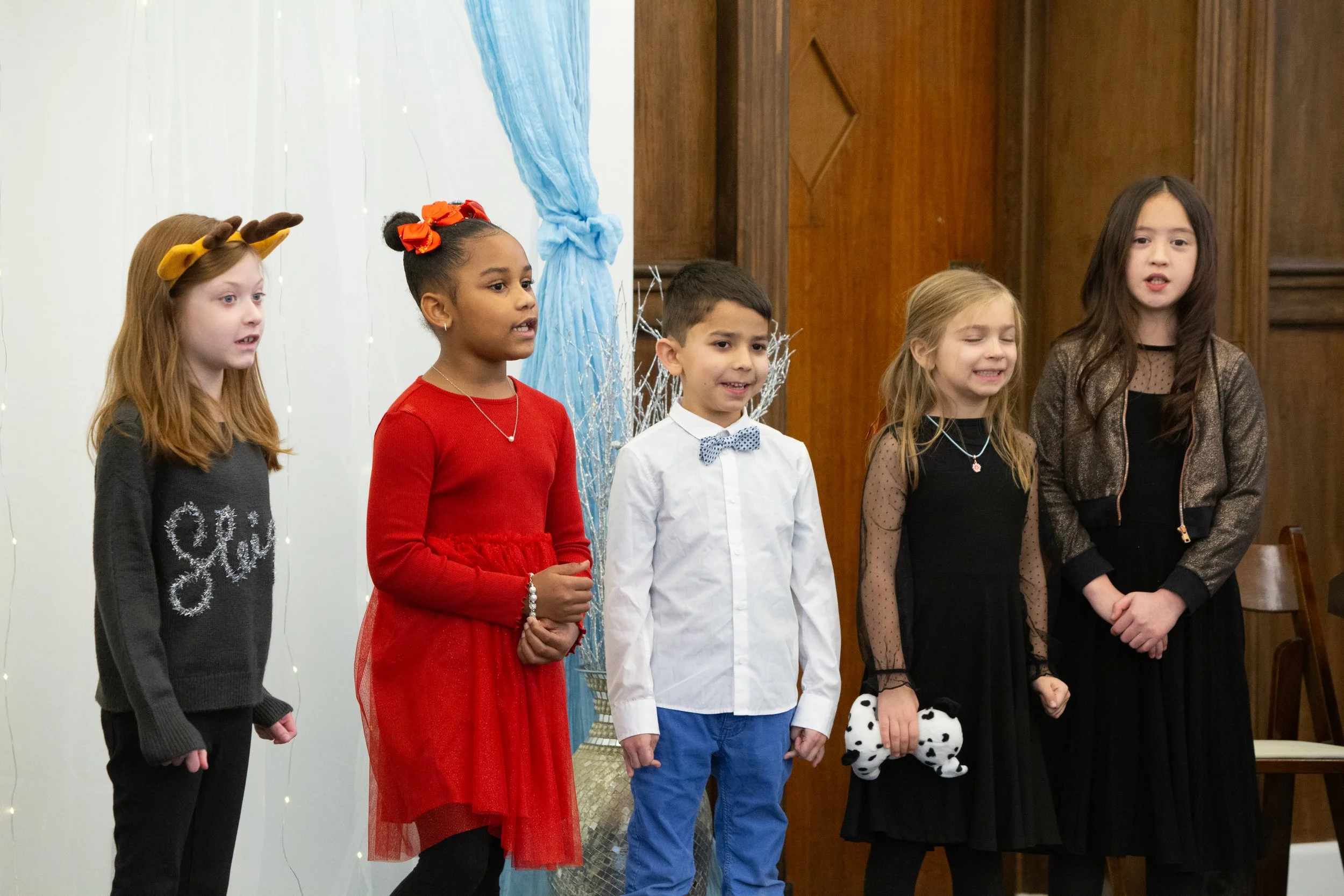 Five children standing in a line, singing or reciting in a Christmas or holiday performance.