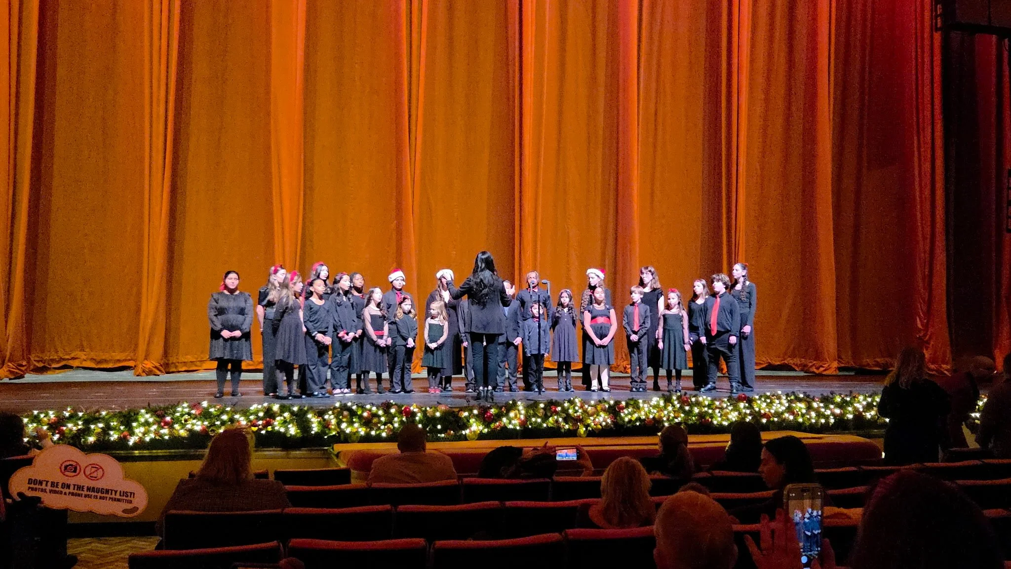 Children's choir performing on stage in a theater with gold curtains, holiday decorations, and Christmas hats.