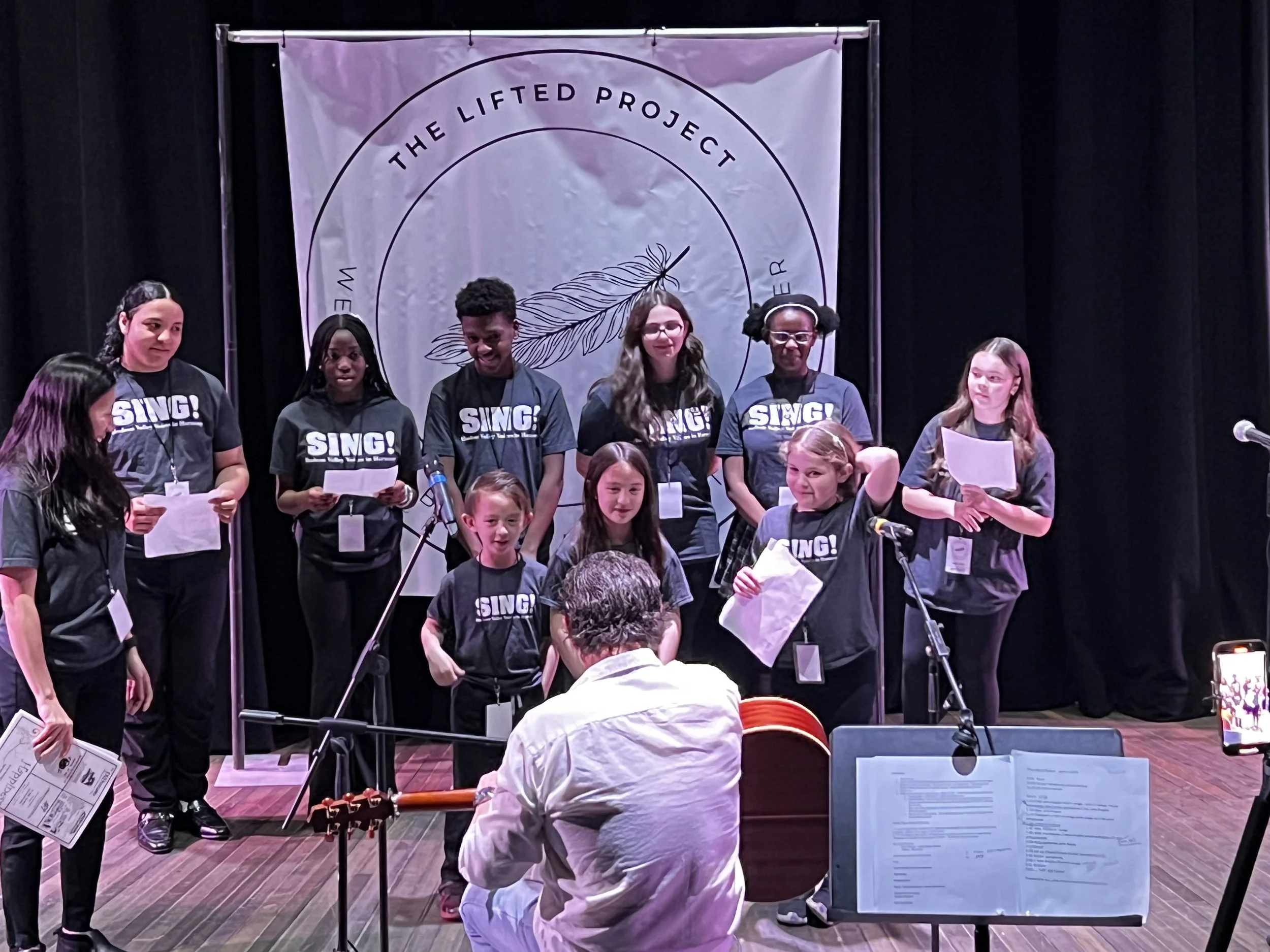 Group of children on stage singing during a performance, with a conductor in front, a backdrop with the words 'The Lifted Project' and a feather logo, microphones, and music stands around them.