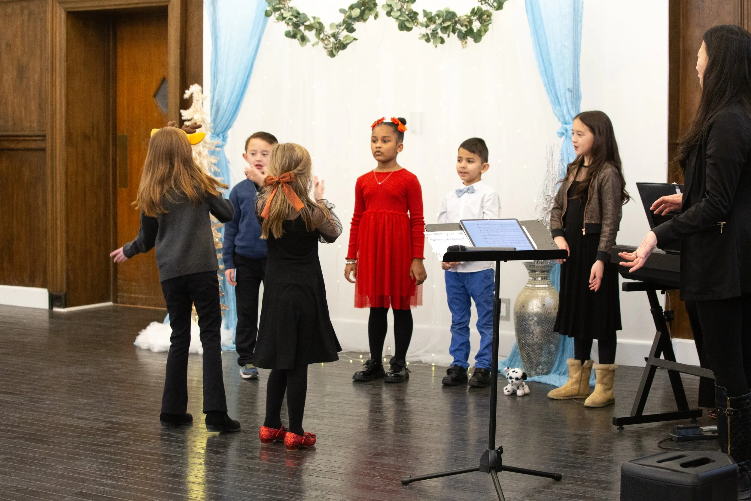 Children performing on stage with a Christmas backdrop, including blue curtains, a white snow-like decoration, and a Christmas tree, with a music stand and a woman playing a keyboard.