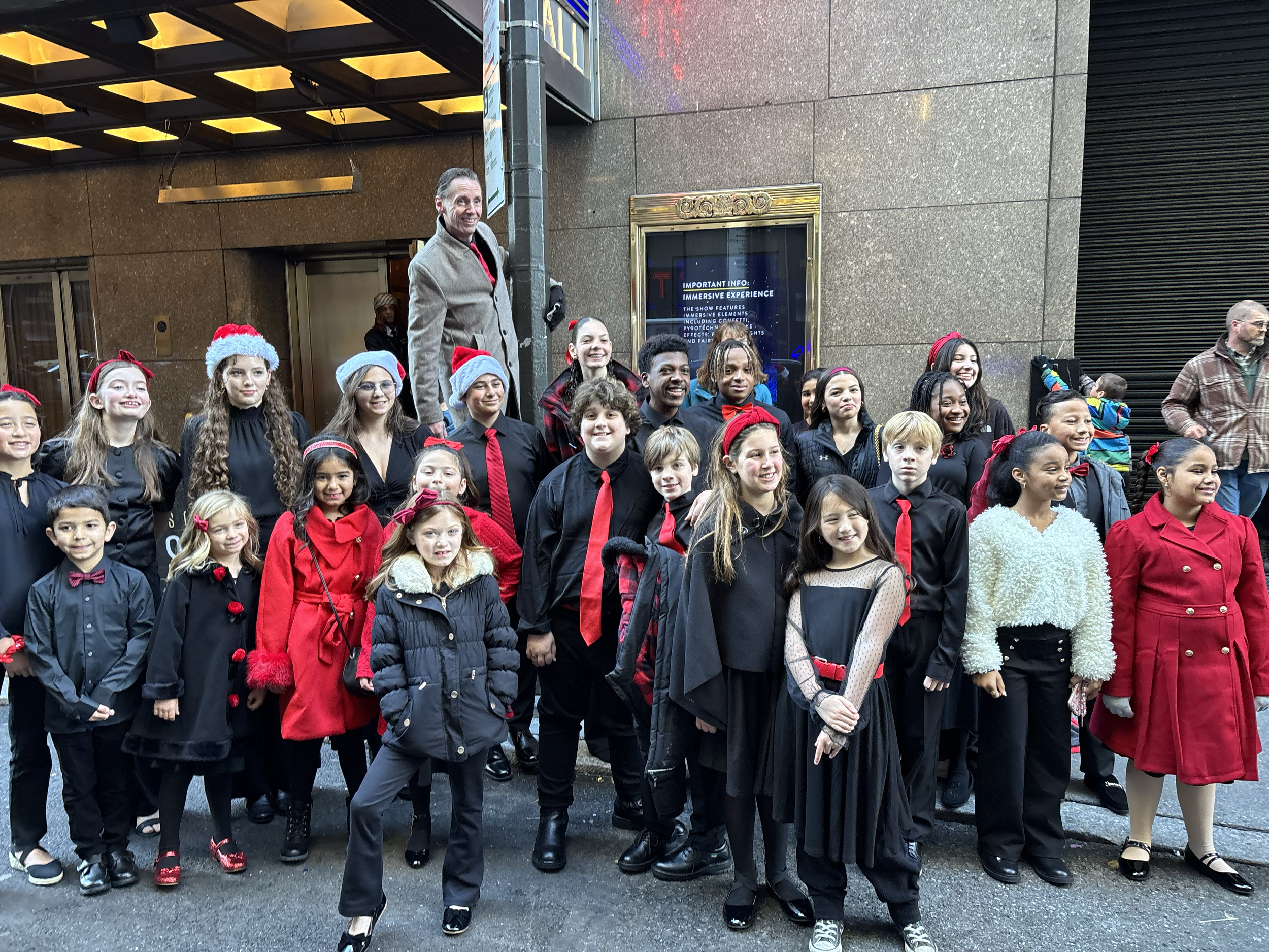 A group of children and adults dressed in black and red holiday attire, including Santa hats and ties, standing on a city sidewalk for a holiday event.
