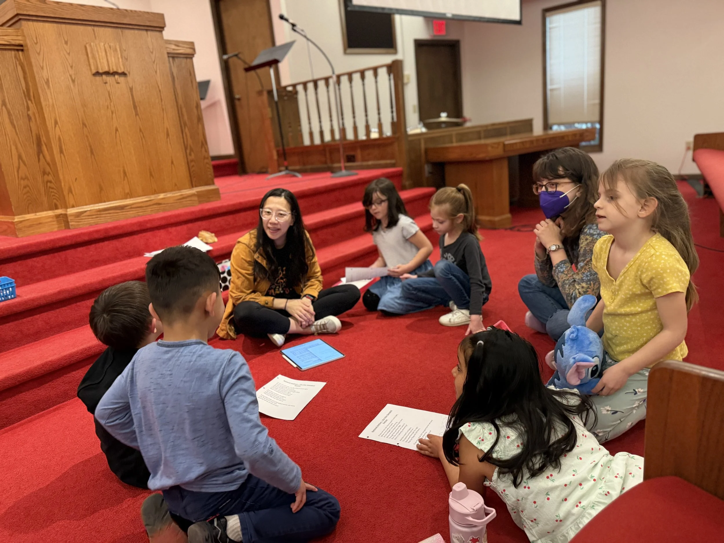 Group of children and two women sitting on red carpeted steps in a church or auditorium, engaging in a discussion or activity, with papers and devices in front of them.