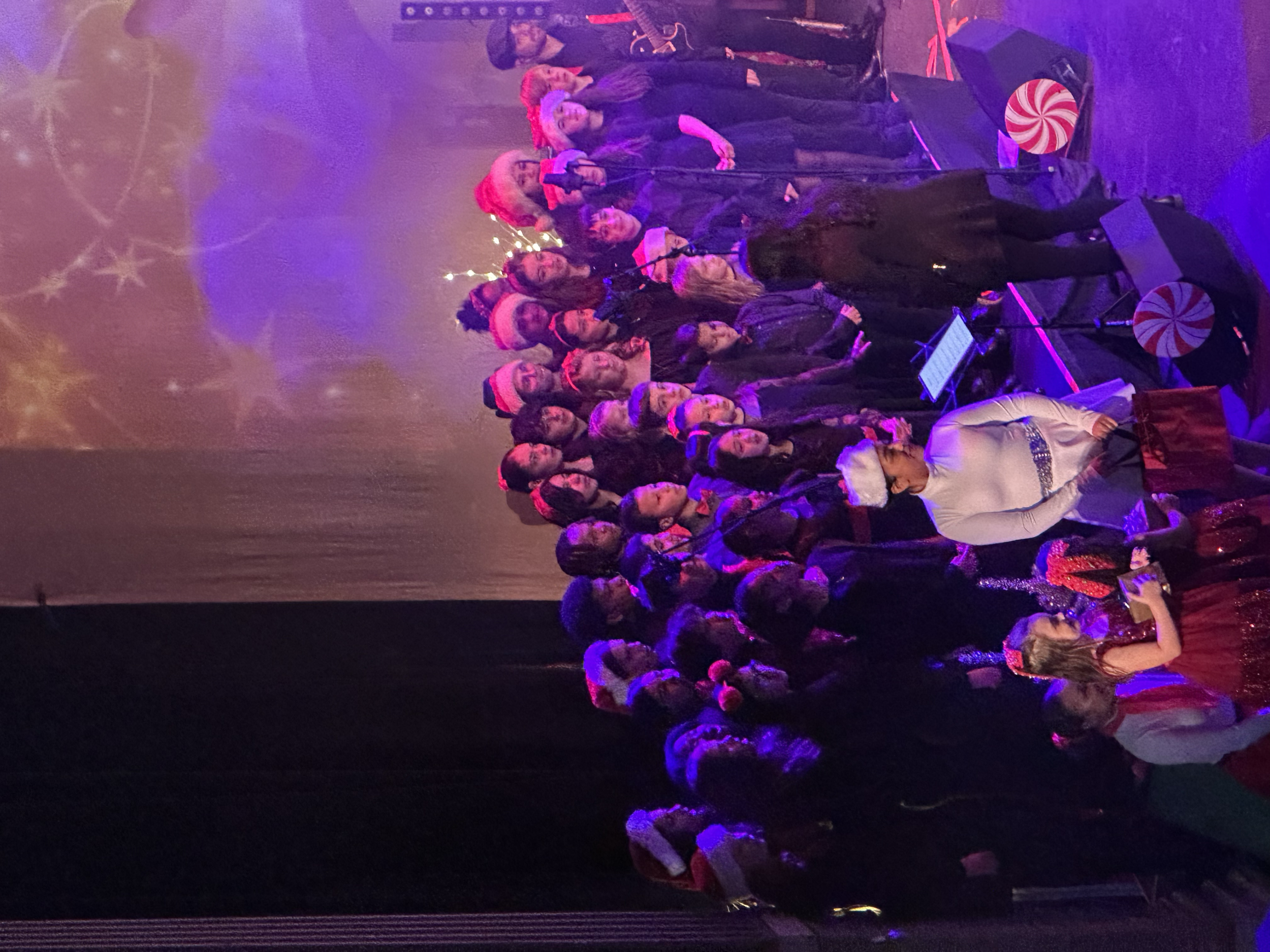 A large group of people celebrating Christmas, many wearing Santa hats, on a decorated stage with Christmas-themed props, including candy canes and a large peppermint ornament, with festive lighting.