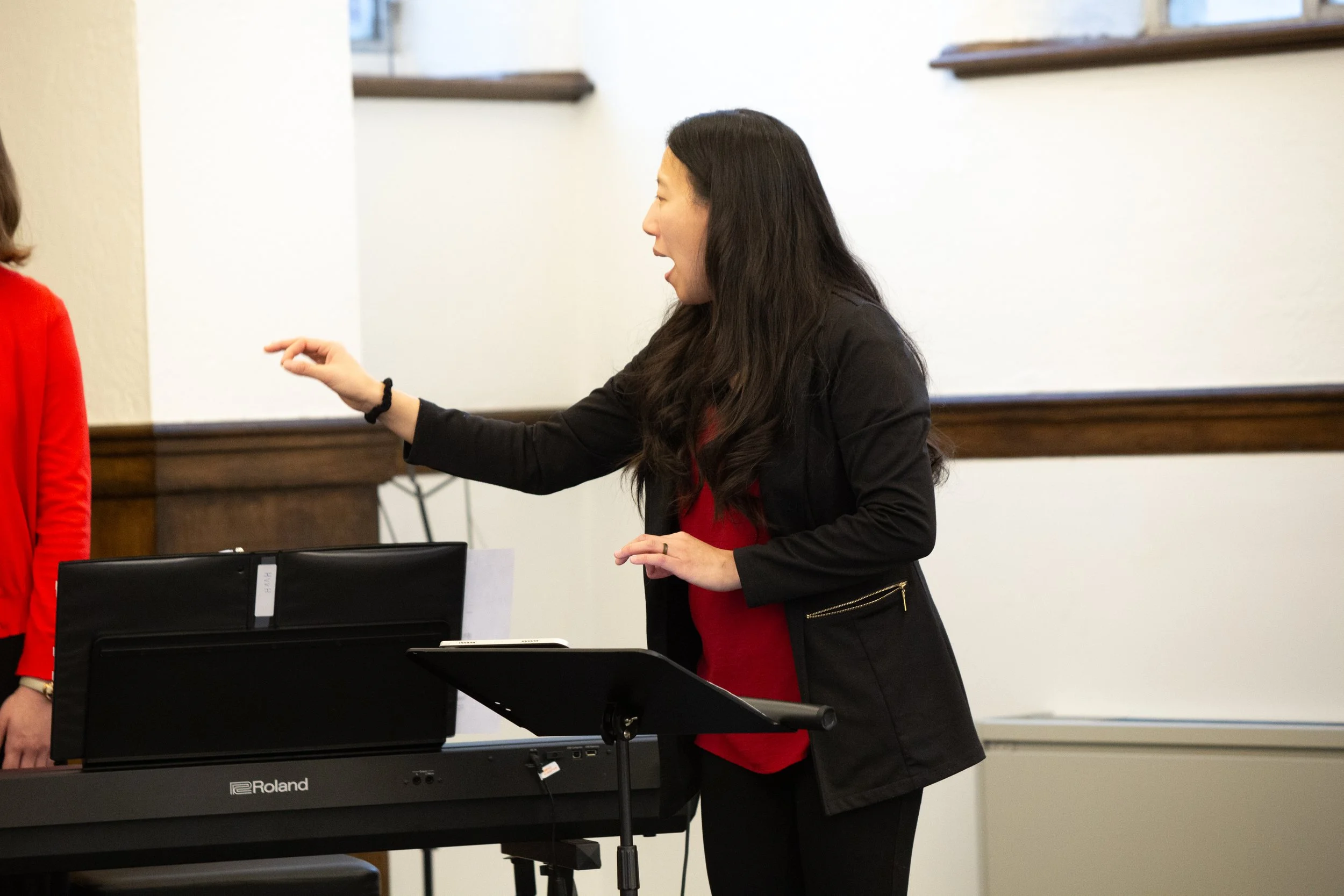 A woman with long dark hair appears to be speaking or explaining something, with her right hand extended with a finger pointing. She is standing next to a digital piano, dressed in a black blazer and red top, in a room with white walls and wood trim.