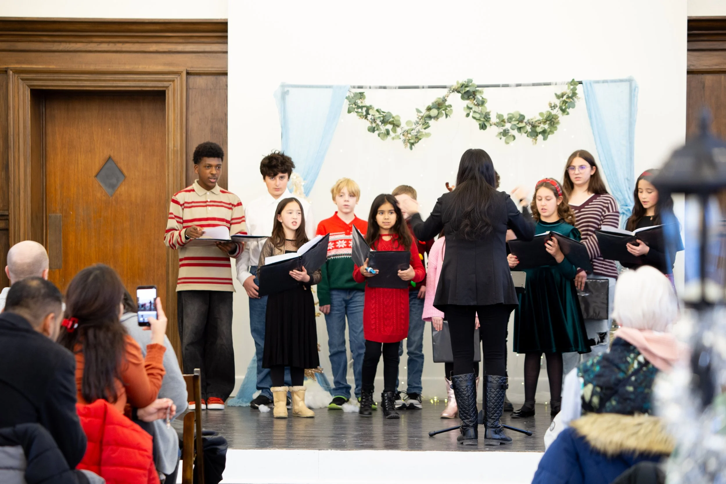 A group of children standing on a stage singing Christmas carols, with a woman conducting them, in front of an audience.
