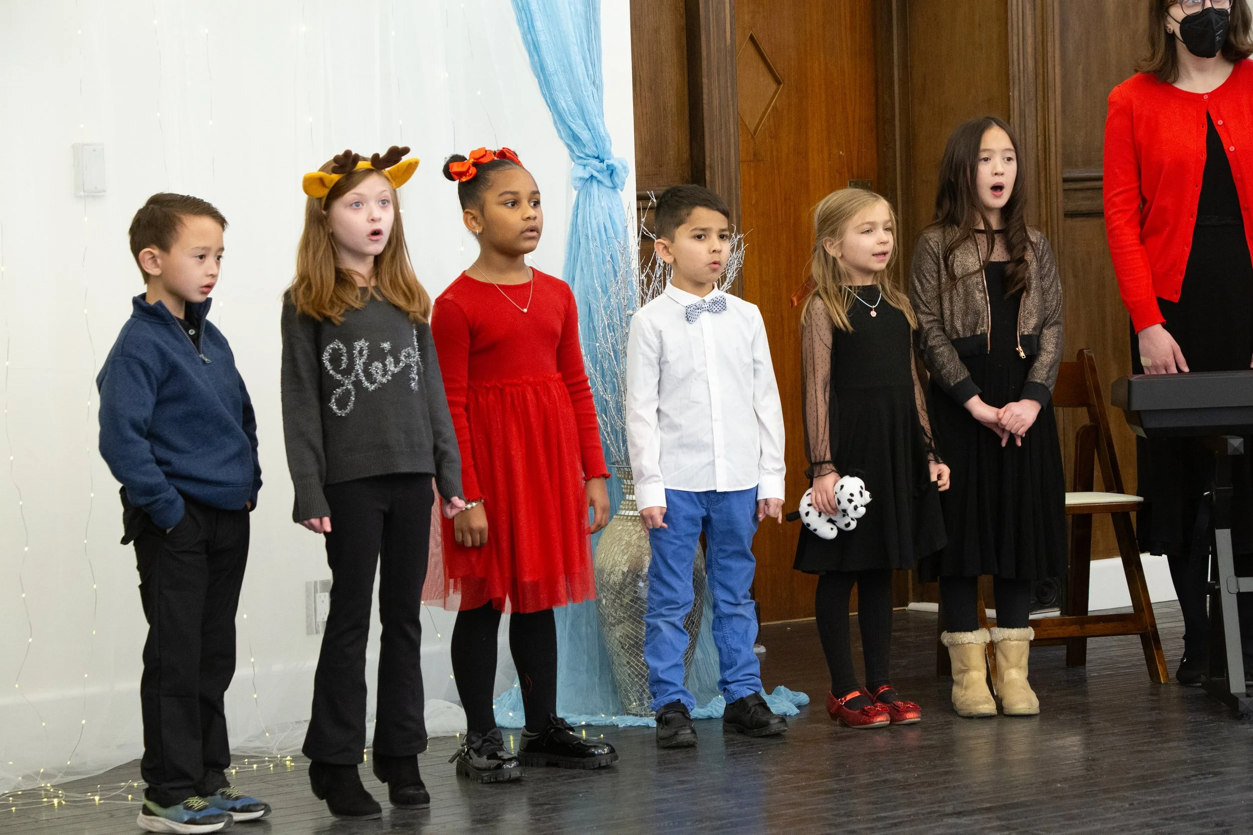 Children singing in a holiday performance, dressed in festive clothing, standing in a row on stage.