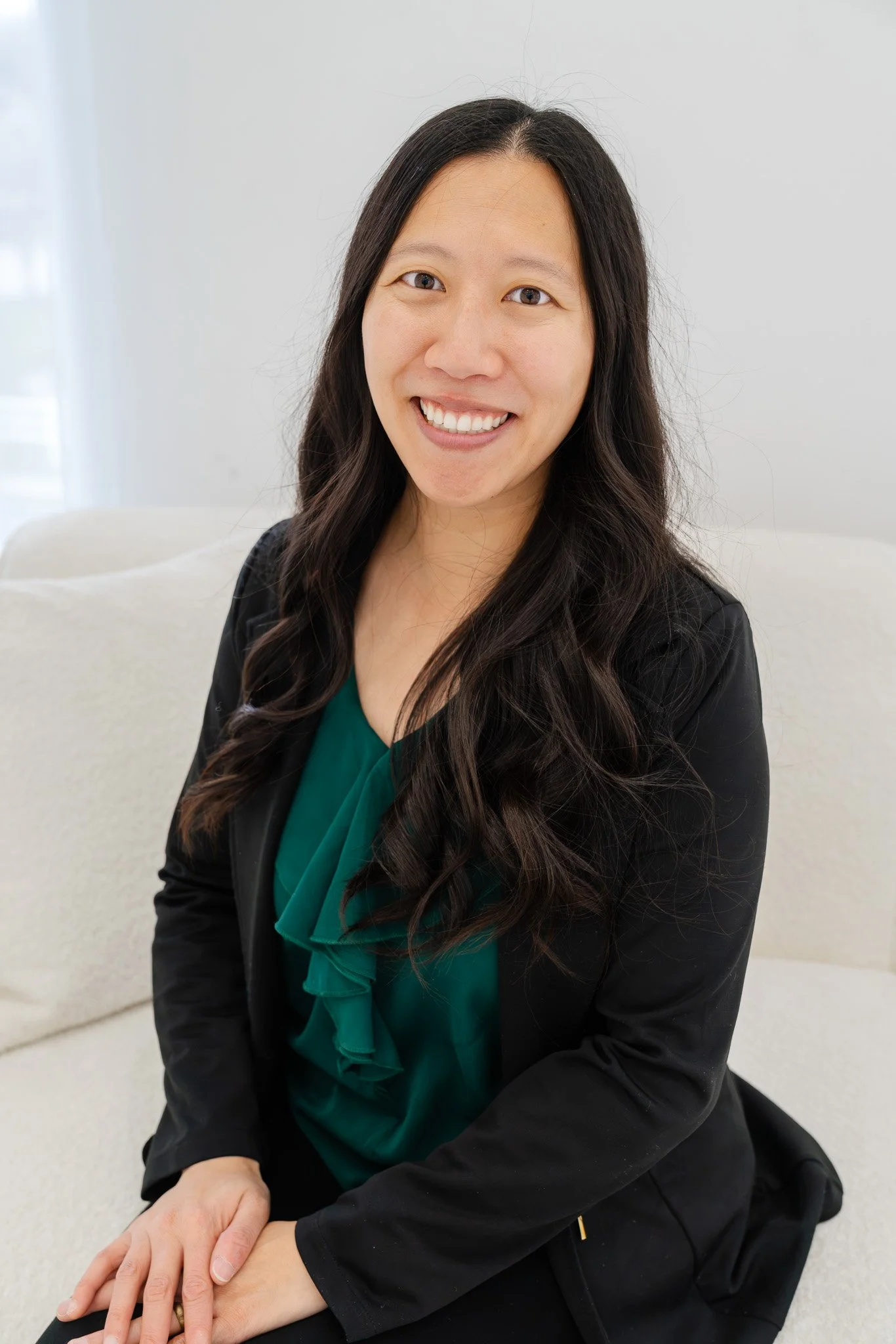 A woman with long dark hair smiling, sitting on a white couch in a well-lit room, wearing a black blazer over a green blouse.