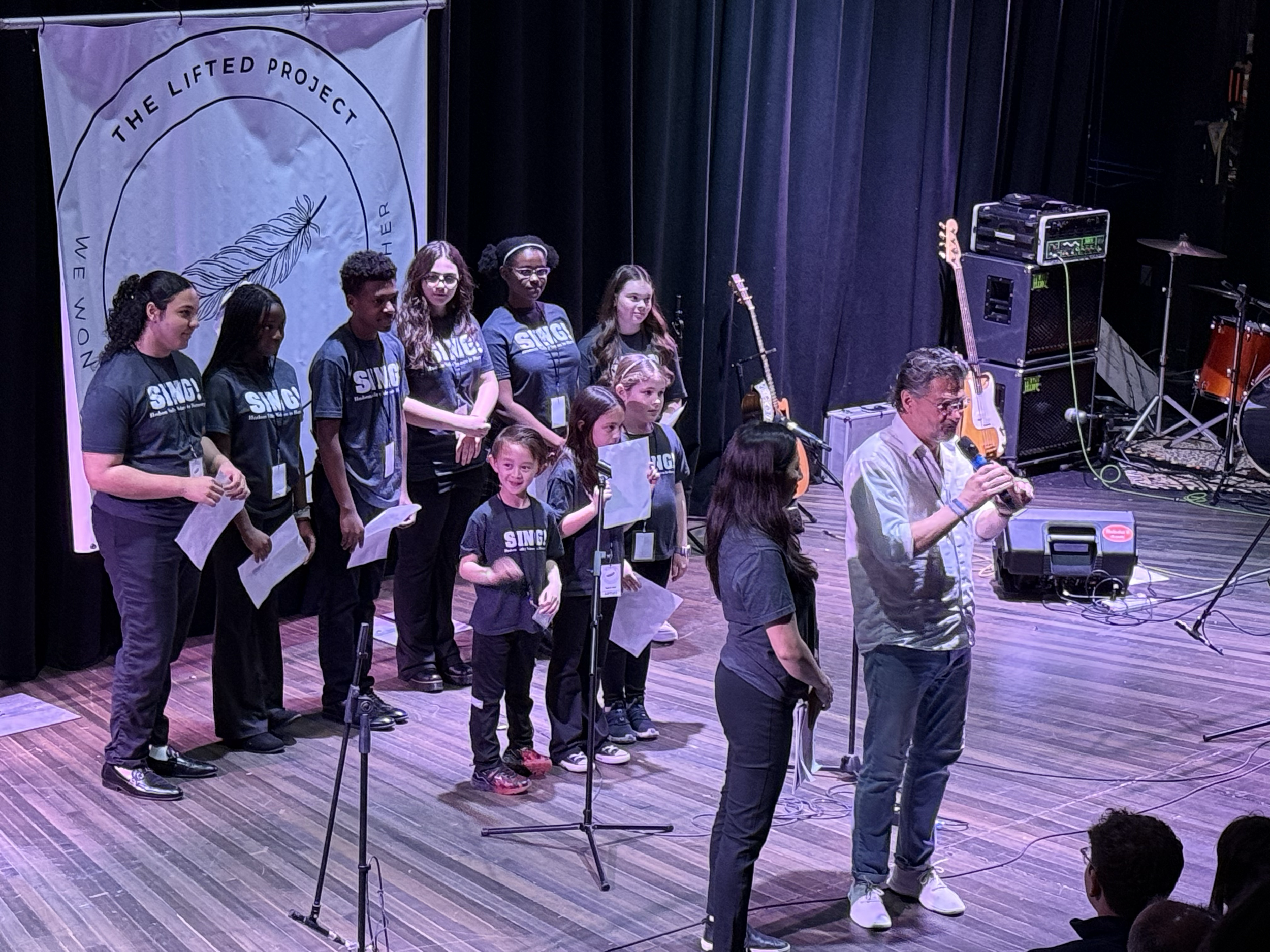 A group of children and a few adults on stage during a singing performance, with musical instruments in the background and a banner that reads 'The Lifted Project' behind them.