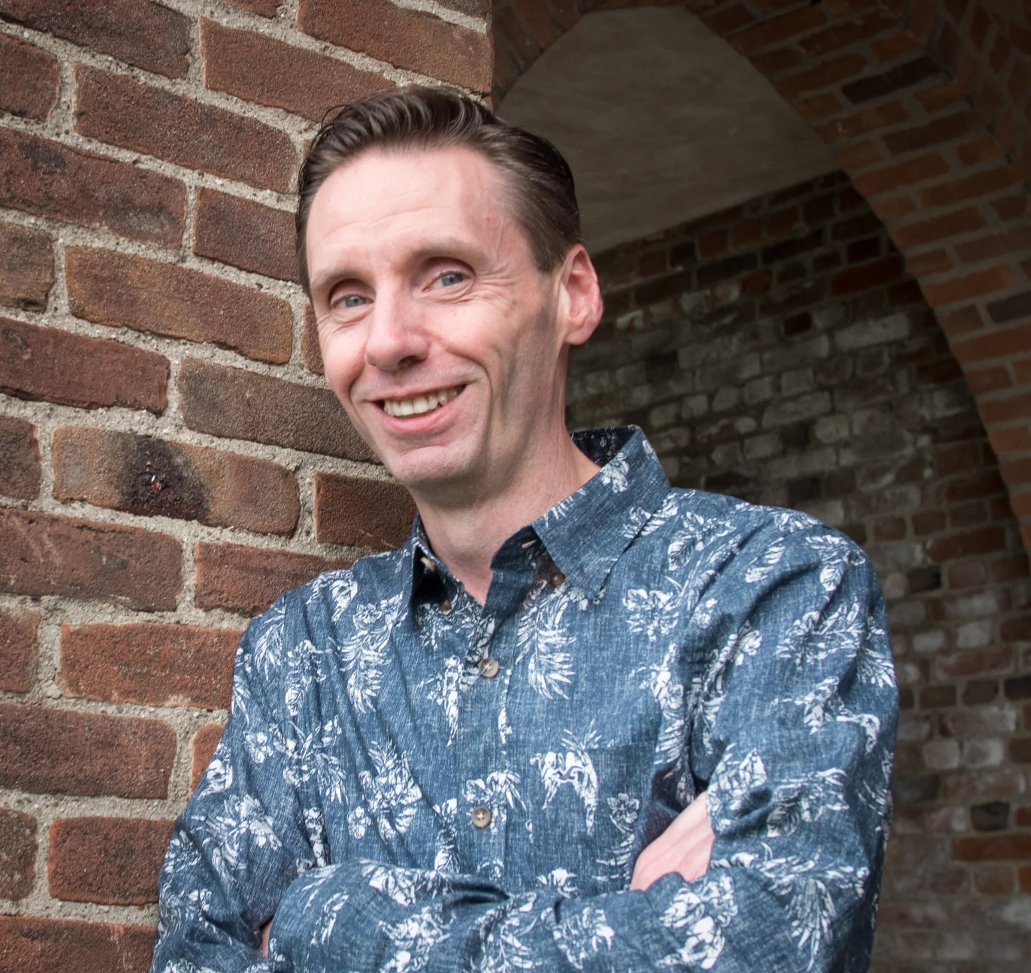 A smiling man with short brown hair wearing a blue patterned shirt, standing in front of a brick wall and arched brick structure.